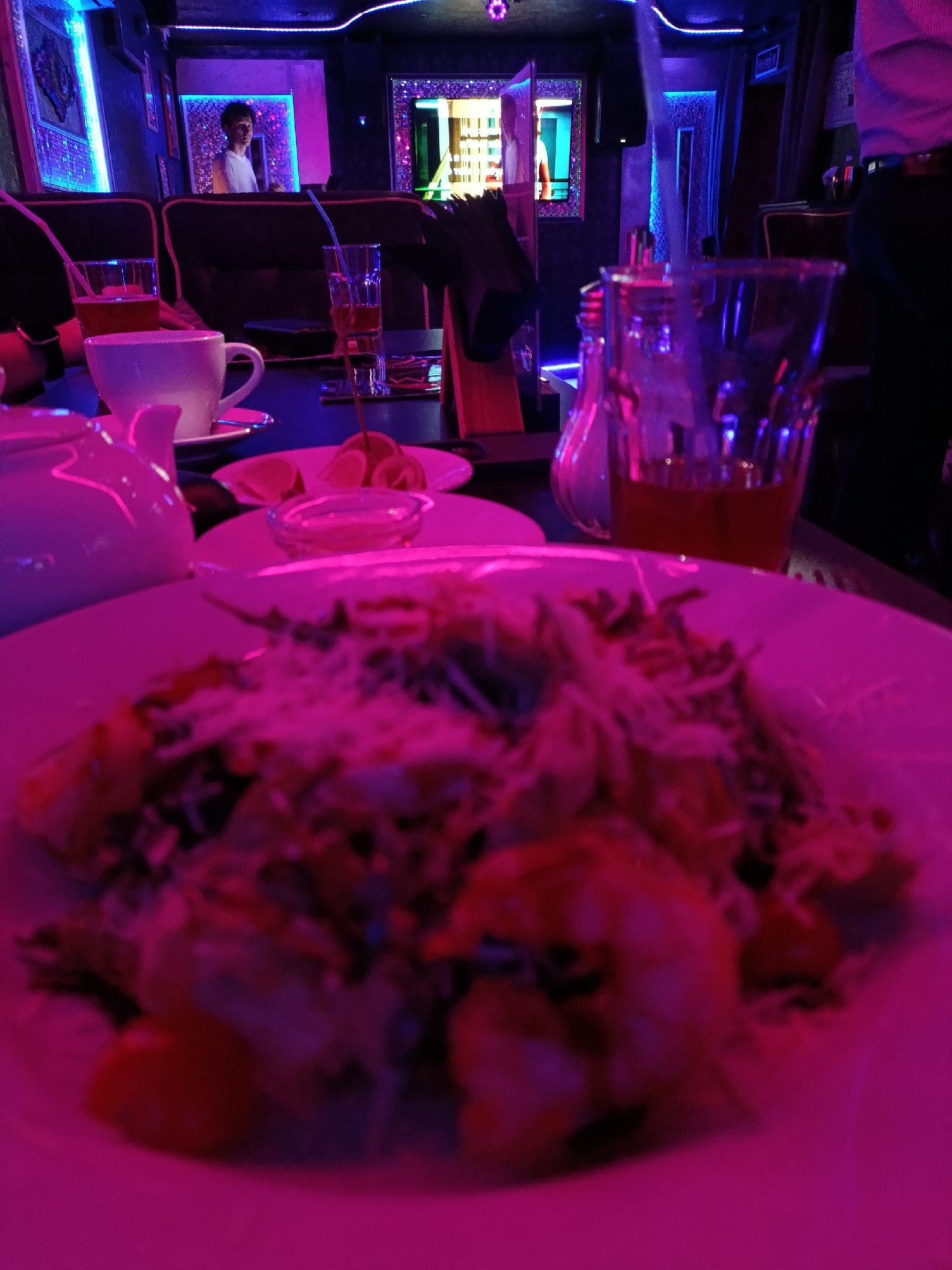 A full-shot, eye-level, medium-distance indoor shot shows a table in a dimly lit, possibly nightclub or lounge setting. The lighting is predominantly pink and blue. In the foreground, a white plate holds a portion of a salad, with visible ingredients like shredded cheese, greens, and what appear to be pieces of shrimp or other seafood. To the left of the plate is a white teapot. Further back on the table, there are two glasses, one containing a dark amber liquid and a straw, and another smaller glass with a pale liquid. A white teacup sits on a saucer. In the background, a young man in a white t-shirt is visible standing near a wall with a large, illuminated mirror. The environment appears to be a public establishment with decorative lighting. The scene is likely in Moscow, Russia, given the context of the query.