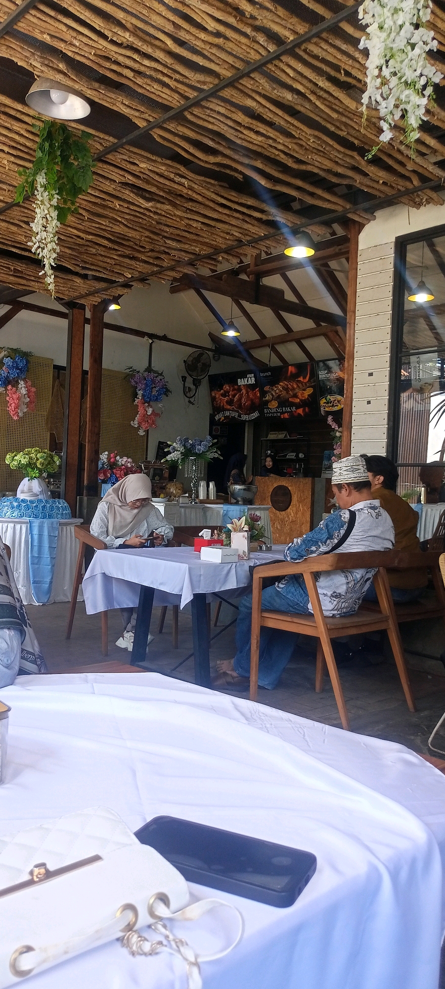 In a casual outdoor dining area with a rustic wooden ceiling structure, two individuals are seated at a table. A woman in a light brown hijab and glasses is looking down at her phone while seated at a table covered with a light purple cloth. Across from her, a man wearing a white and blue patterned shirt and a white skullcap is seated on a wooden chair, his back to the camera. He is looking down and his legs are visible. Another person, partially visible and wearing a brown shirt, is seated to the right of the man. The table is set with white boxes and what appear to be flowers in vases. In the background, a counter area is visible with a digital menu board displaying images of grilled food. The floor is paved. The location is identified as Desa Ponokawan, Indonesia.