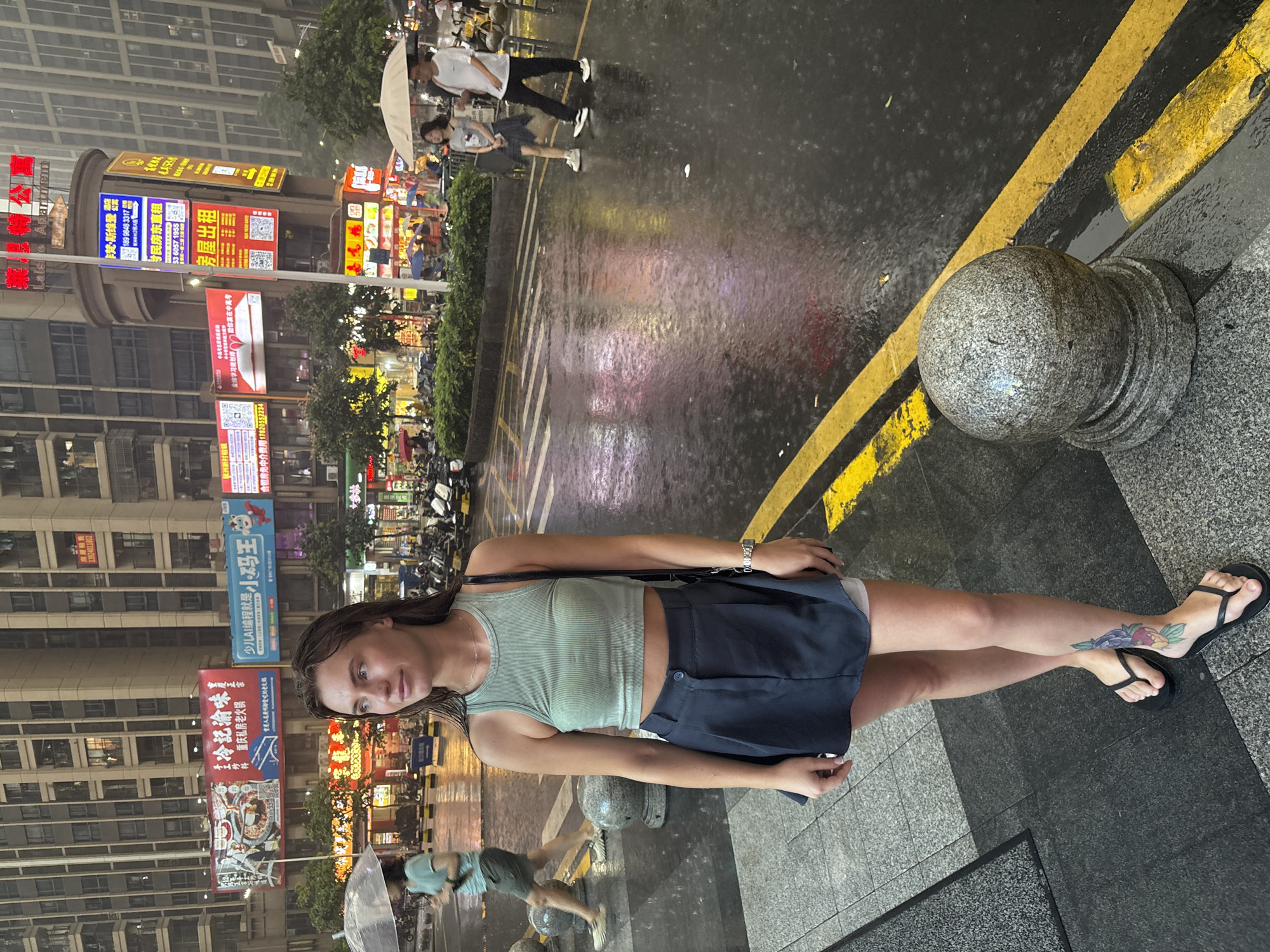 In the city of Haizhu District, China, a young Caucasian woman with light brown hair is standing on a wet sidewalk next to a street. She is wearing a green cropped tank top, a dark blue pleated skirt, and black flip-flops. She has a tattoo of a flower on her left leg. The street is wet with rain, reflecting the lights of the surrounding buildings and advertisements. Several people are walking in the background, some holding umbrellas. Motorcycles are parked along the side of the street. The buildings are tall and modern, with many windows and illuminated signs. The sky is overcast, indicating it is raining.