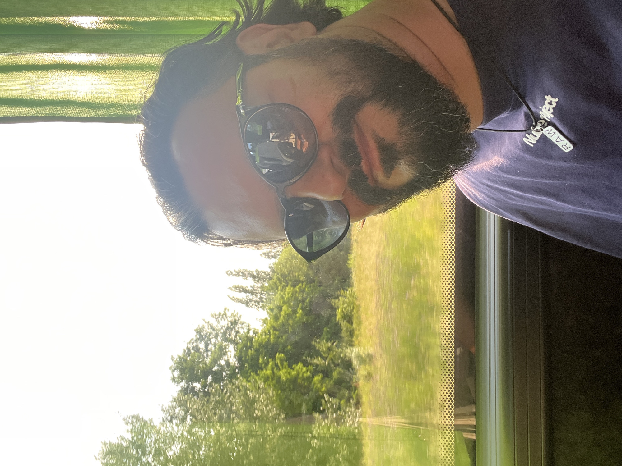 A man with dark hair and a beard, wearing sunglasses and a dark blue t-shirt, is taking a selfie from inside a vehicle. He is looking towards the camera. Through the window to his left, a blurry green landscape with trees is visible, suggesting motion. To his right, a bright green curtain is drawn, with sunlight filtering through it. The man is wearing a necklace with two pendants. One pendant appears to be a rectangular tag with the word "RAW" on it, and the other is a stylized word that reads "Nuproject". The scene appears to be from a sunny day, likely during daylight hours. There is no indication of the specific location within Antwerp, Belgium.