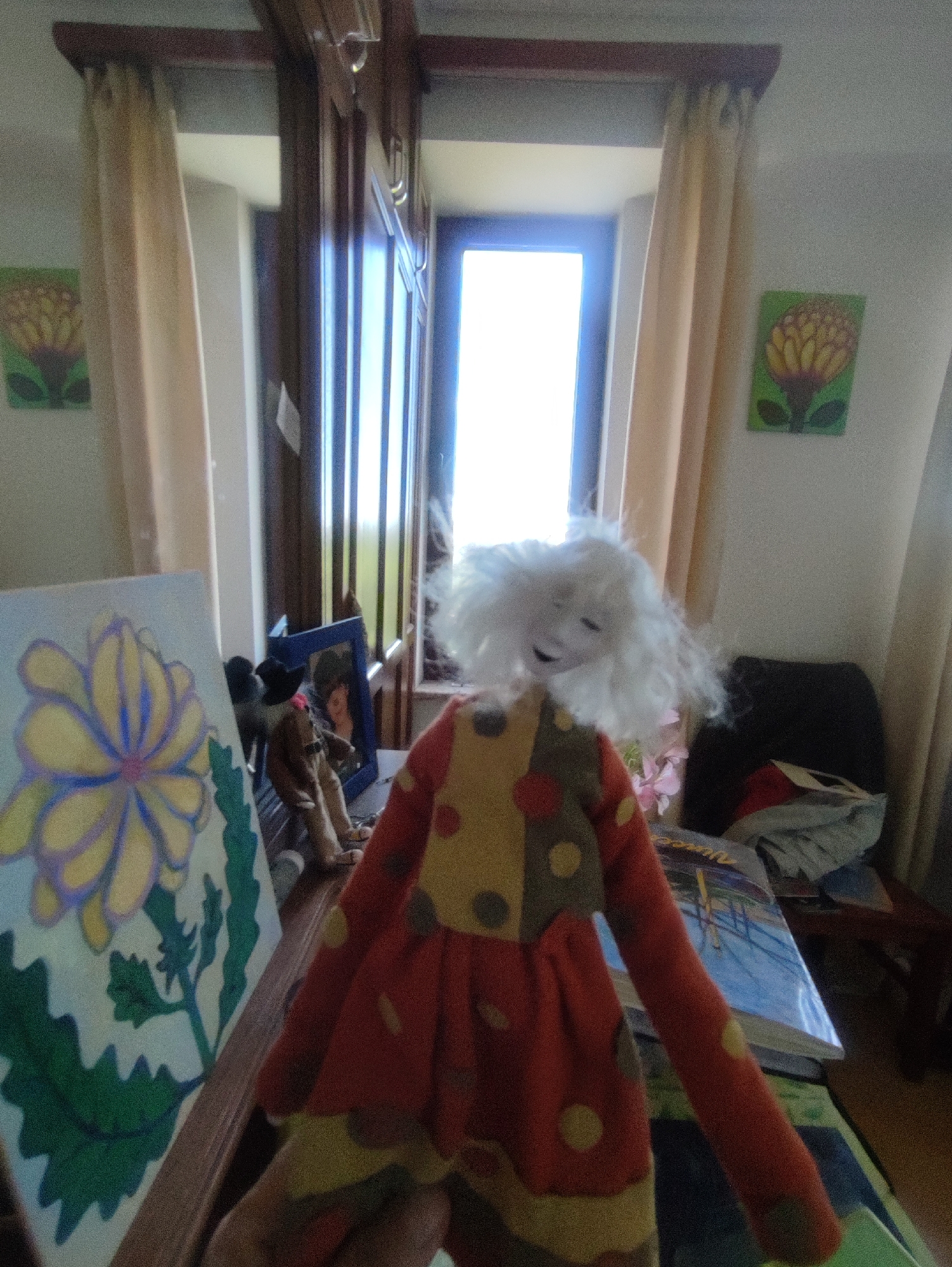 The image shows a room interior. A handmade doll with white hair and a red and yellow polka-dotted outfit is in the foreground. To the left, there's a painting of a yellow flower on a blue canvas. In the background, there is a window with light shining through, flanked by beige curtains. On the walls, there are two flower paintings, and there's a dark chair in the background. A wooden framed picture and a small doll also rest on a table. The city of Yerevan, Armenia, is not discernible from the image, and the description is based solely on the visual content.