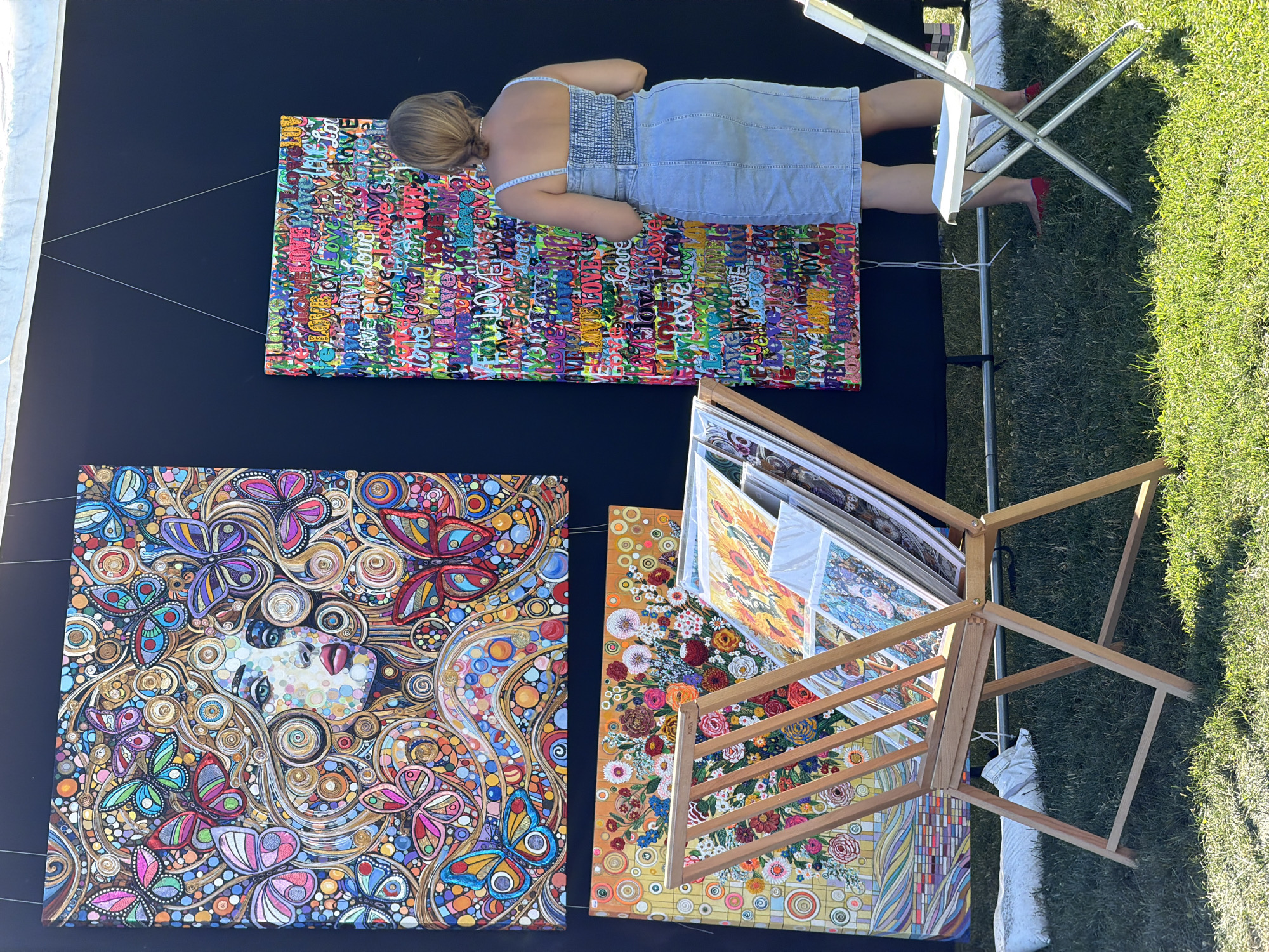 This image captures an outdoor art exhibition, likely in a village setting, as suggested by the presence of a tent structure and lush green grass. The foreground is dominated by a wooden display rack filled with various framed artworks, suggesting a focus on visual arts. In the midground, three large, vibrant paintings are displayed against a black backdrop. The leftmost painting is a detailed mosaic-like portrait of a woman surrounded by colorful butterflies and intricate patterns. To its right is a tall, narrow painting composed of numerous repetitions of the word "love" in various fonts and colors. Below these, a horizontally oriented painting depicts a floral arrangement with a yellow background. A woman, dressed in a denim sundress and red flats, is visible in the right portion of the image, observing the artwork. Her stance and focus indicate engagement with the art. The environment appears to be a sunny day, with dappled light on the grass, and a white folding chair is positioned to the right, adding to the outdoor exhibition feel. The overall scene suggests a community event or art fair, possibly within the Village of Sag Harbor, United States of America, given the mention of a village context.