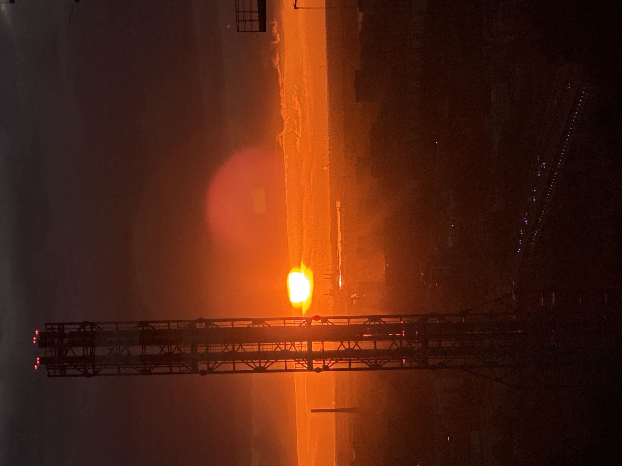The image depicts a sunset observed in the city of Unknown, Unknown. In the foreground is a steel lattice tower. The sun is positioned near the horizon, visible through the tower's structure. The sky is obscured by clouds and haze, colored in shades of orange and red. To the right, the city lights are scattered.