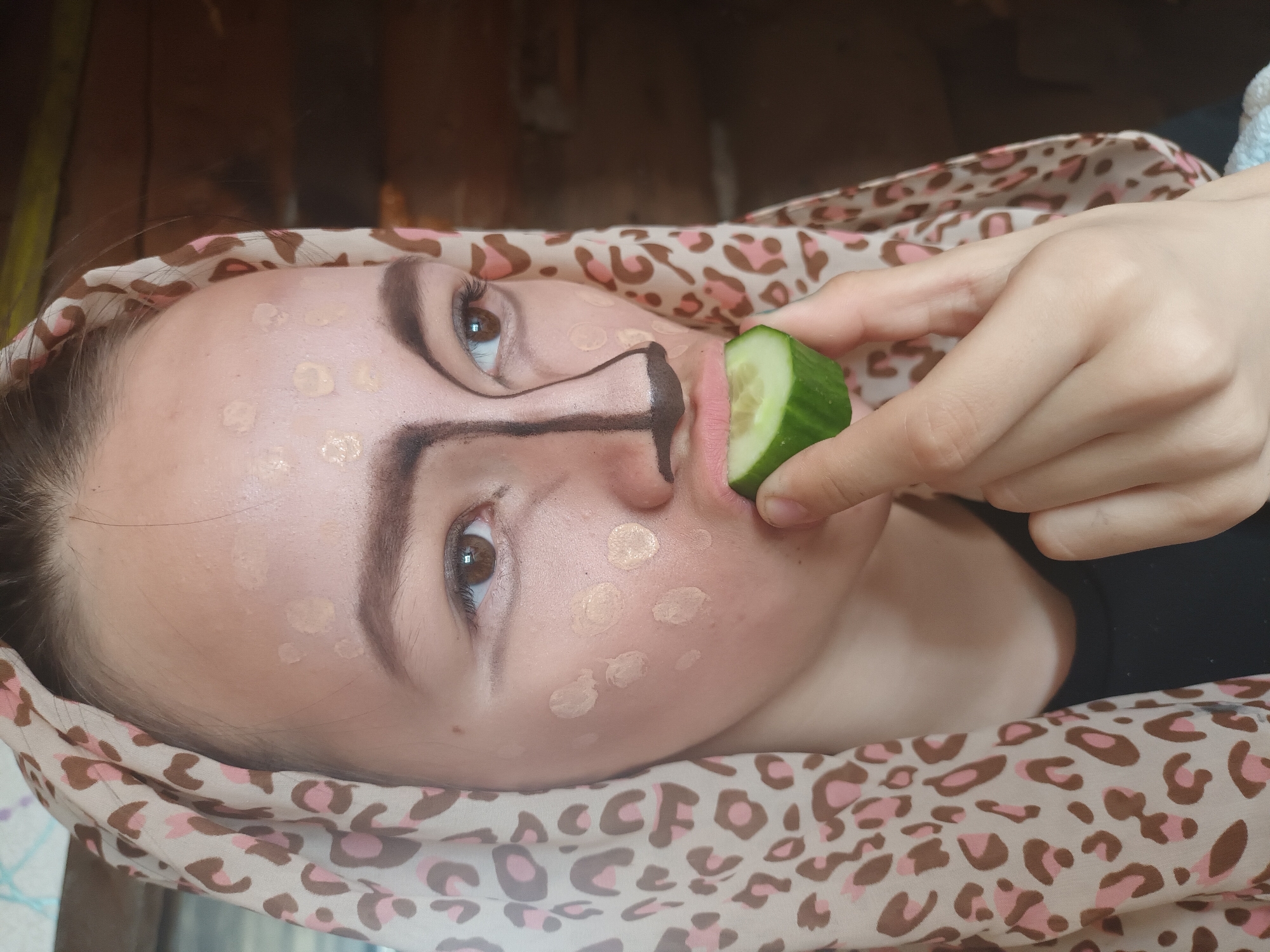 A close-up, high-angle portrait captures a young person, likely in Ольховка, Russia, engaged in applying theatrical or costume makeup. The subject is positioned horizontally, with their head resting on a surface, gazing directly at the viewer.
Their face is adorned with numerous dots of light-toned foundation or concealer, indicating an incomplete makeup application. Prominent dark brown or black lines are drawn to create the distinctive features of an animal's nose and mouth, suggesting a feline or similar creature. The subject has well-defined eyebrows and subtle eye makeup, enhancing their gaze.
In their right hand, they delicately hold a fresh, green slice of cucumber, positioning it playfully near their lips. This adds a whimsical element to the scene, possibly as a prop or a break from the makeup process.
A light-colored fabric, featuring a distinctive pink and brown leopard print pattern, is draped around the person's head and neck, serving as a stylish headscarf or wrap. The background is indistinct and dark, suggesting an indoor setting.
The image conveys a sense of creative exploration and playful self-transformation. No text is visible in the frame.