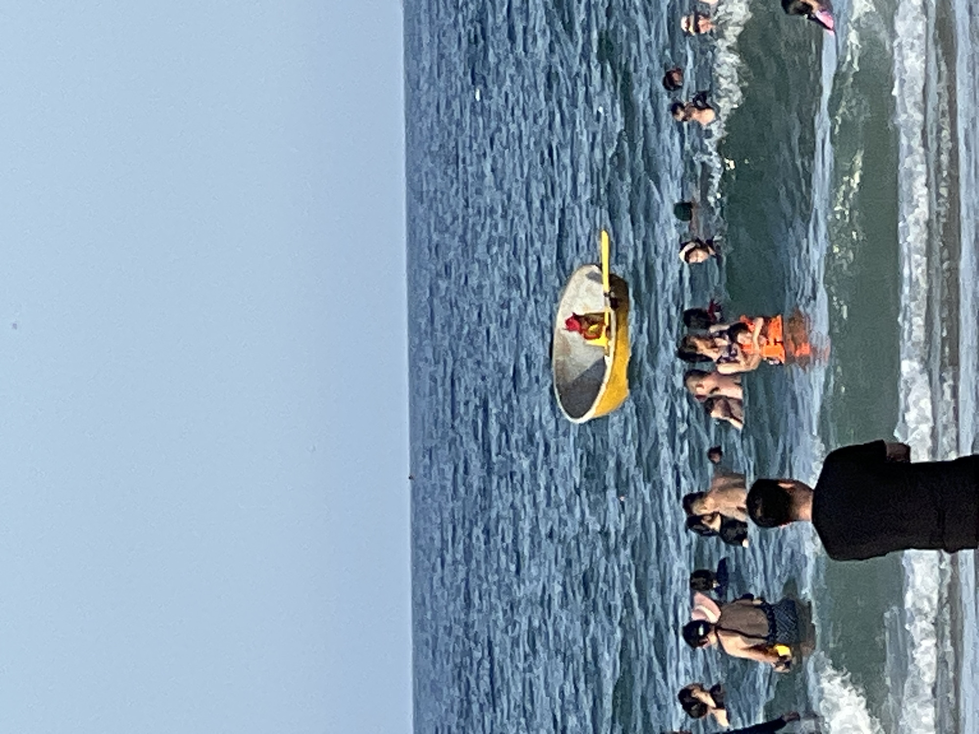A group of people are in the ocean on a sunny day. In the middle of the frame, a small yellow boat with a person in a red hat and orange life jacket is visible. Several people are swimming and playing in the water around the boat. The ocean is a deep blue, with gentle waves. The sky is a pale blue and clear. The scene is likely at a beach in Da Nang, Vietnam, a popular tourist destination known for its coastline and watersports.