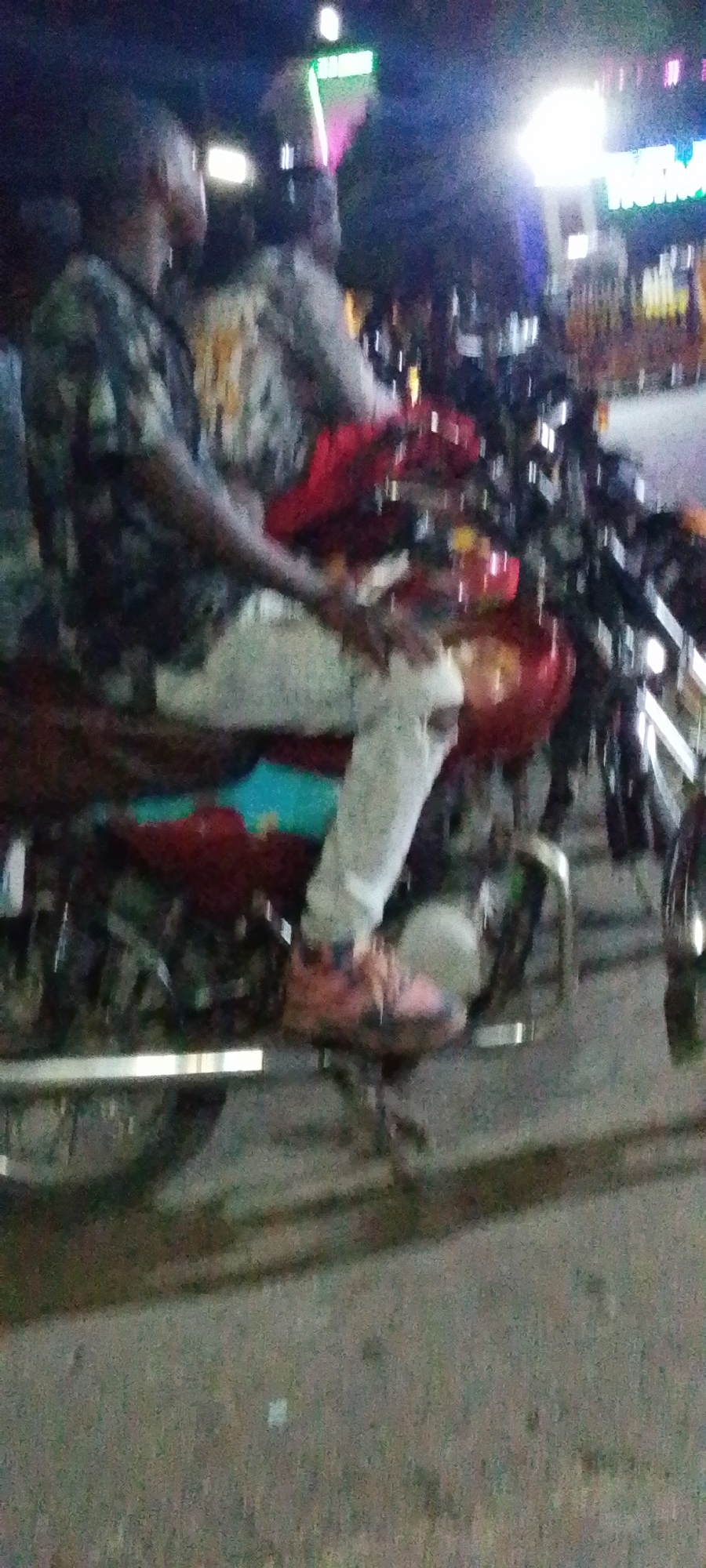 The image shows two men on a red motorcycle, parked on a street in Lokoja, Nigeria. The man in the foreground is wearing a dark, patterned shirt and light-colored pants. He has pink sneakers on his feet. Another man is sitting behind him, with a grey shirt. Several other motorcycles are parked alongside. Streetlights and bright signs illuminate the background, with trees present at the top of the image. The foreground is asphalt.