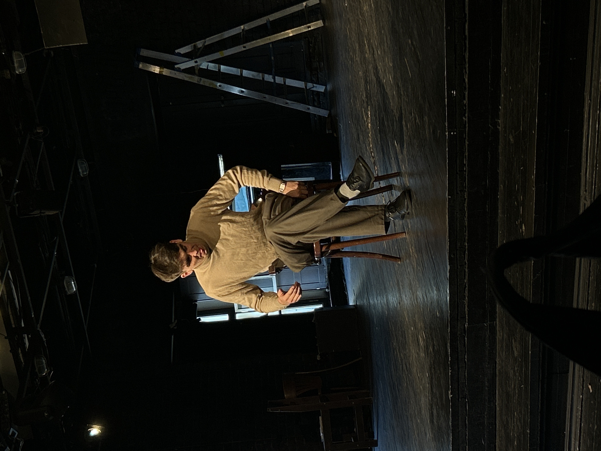 A man is seated on a wooden chair on what appears to be a stage or rehearsal area. He wears a light-colored long-sleeved top, possibly a turtleneck, and light brown or olive-green trousers. A watch is visible on his left wrist. He leans slightly forward, with his hands raised and open, suggesting he is performing, speaking, or deep in thought. His expression is focused and serious.
The floor beneath him is composed of dark, possibly painted, wooden floorboards. The background is dimly lit and dark, featuring several metal ladders propped against a dark wall or structure. Industrial-style lights or stage fixtures are sparsely distributed in the darkness, some with visible clear glass covers. To the man's right, a dark wooden structure, possibly a table or prop, is also visible. The artificial, focused lighting highlights the man against a dark background, creating a theatrical atmosphere. The scene evokes an indoor setting, likely a theater or studio, possibly during a rehearsal or performance. No text is visible in the image.