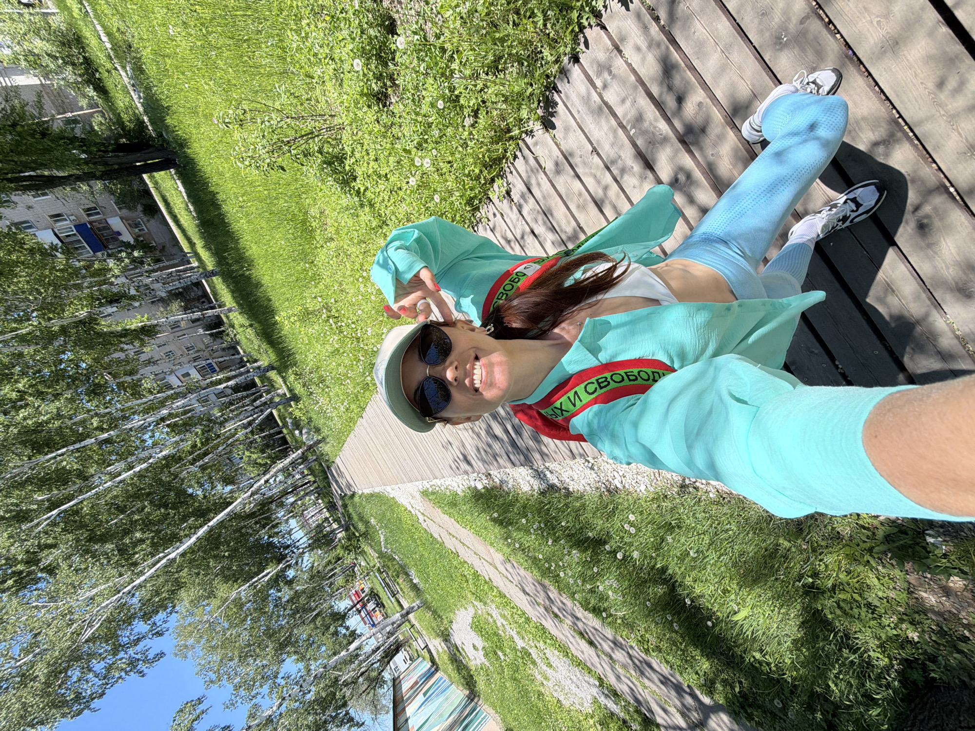 A woman takes a selfie on a wooden boardwalk next to a grassy area with birch trees and apartment buildings. She is wearing a turquoise shirt, a white sports bra, light blue leggings, and white sneakers. A red and green strap is visible across her chest. The sun is shining, and the sky is clear blue. The environment suggests a park or recreational area within a city.