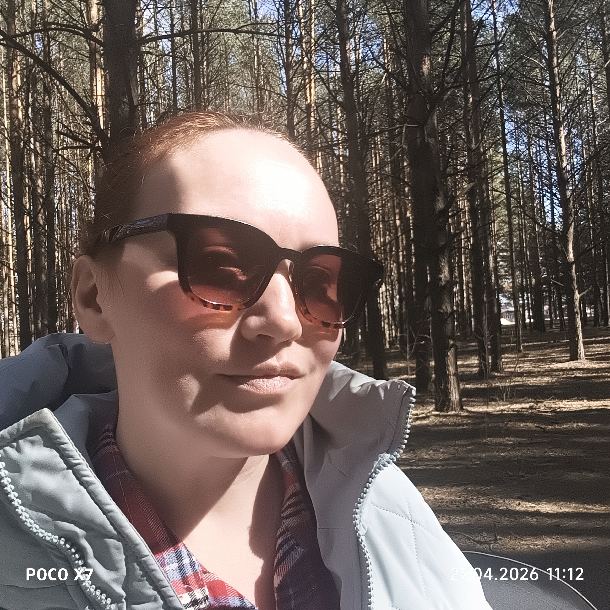 The image displays a well-lit selfie featuring a person, likely an adult woman, positioned in the foreground against a backdrop of a dense forest. She is wearing large, dark, tortoise-shell sunglasses that reflect the surroundings. Her attire includes a light blue, quilted zippered jacket over a red and blue plaid shirt. Strong, direct sunlight illuminates her face, casting notable shadows, suggesting a bright and sunny day. Her expression is largely obscured by the sunglasses but appears neutral or subtly content.
The background is dominated by numerous tall, slender trees, characteristic of a pine or conifer forest, with sunlight filtering through their branches and creating a dappled effect on the forest floor. Patches of blue sky are visible through the tree canopy.
Visible text in the bottom left corner identifies the device as "POCO X7". In the bottom right corner, a timestamp reads "27.04.2026 11:12", indicating the photograph was taken on April 27, 2026, at 11:12 AM. The setting is consistent with forests found in regions like Chaikovsky, Russia.