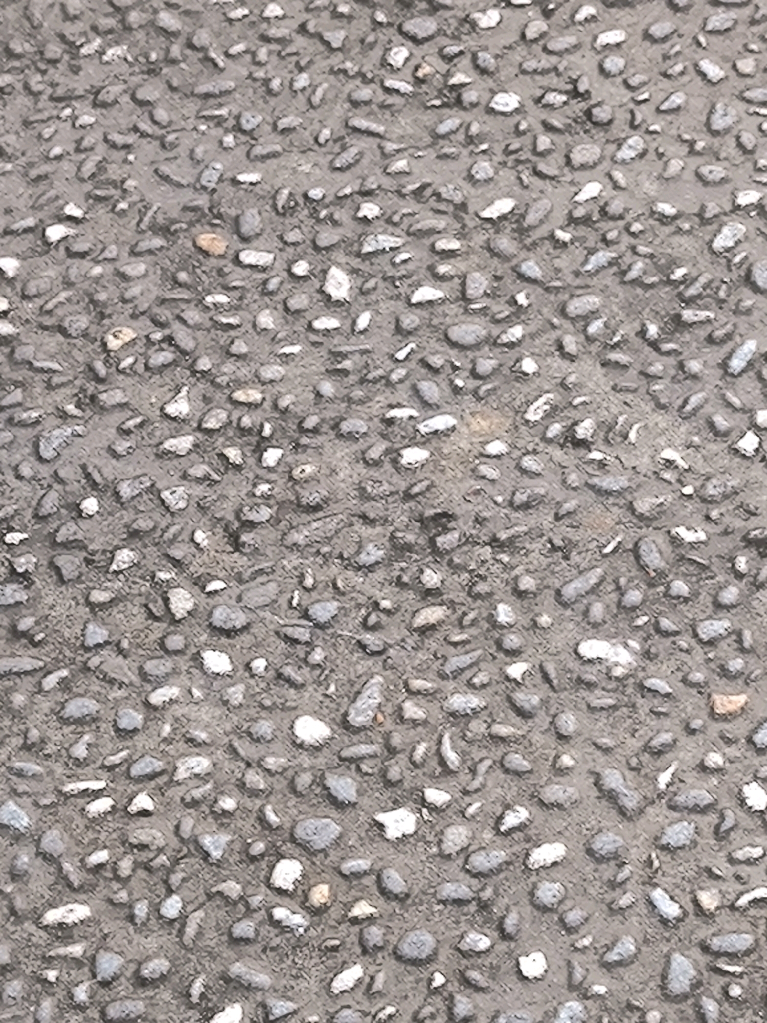 The image shows a close-up, overhead view of coarse asphalt, commonly known as chip seal or tar and chip pavement. The surface is composed of small, irregularly shaped pebbles and stones embedded in a dark gray bituminous binder. The pebbles vary in size, with most ranging from 0.5 to 2 centimeters in diameter. They are predominantly shades of gray, white, and some reddish-brown, creating a speckled pattern. The binder appears slightly wet or worn, giving the surface a somewhat uneven texture. The image lacks any discernible context of location within a city, or any signs of people, specific subjects, objects, or actions, making it a generic depiction of a pavement surface.