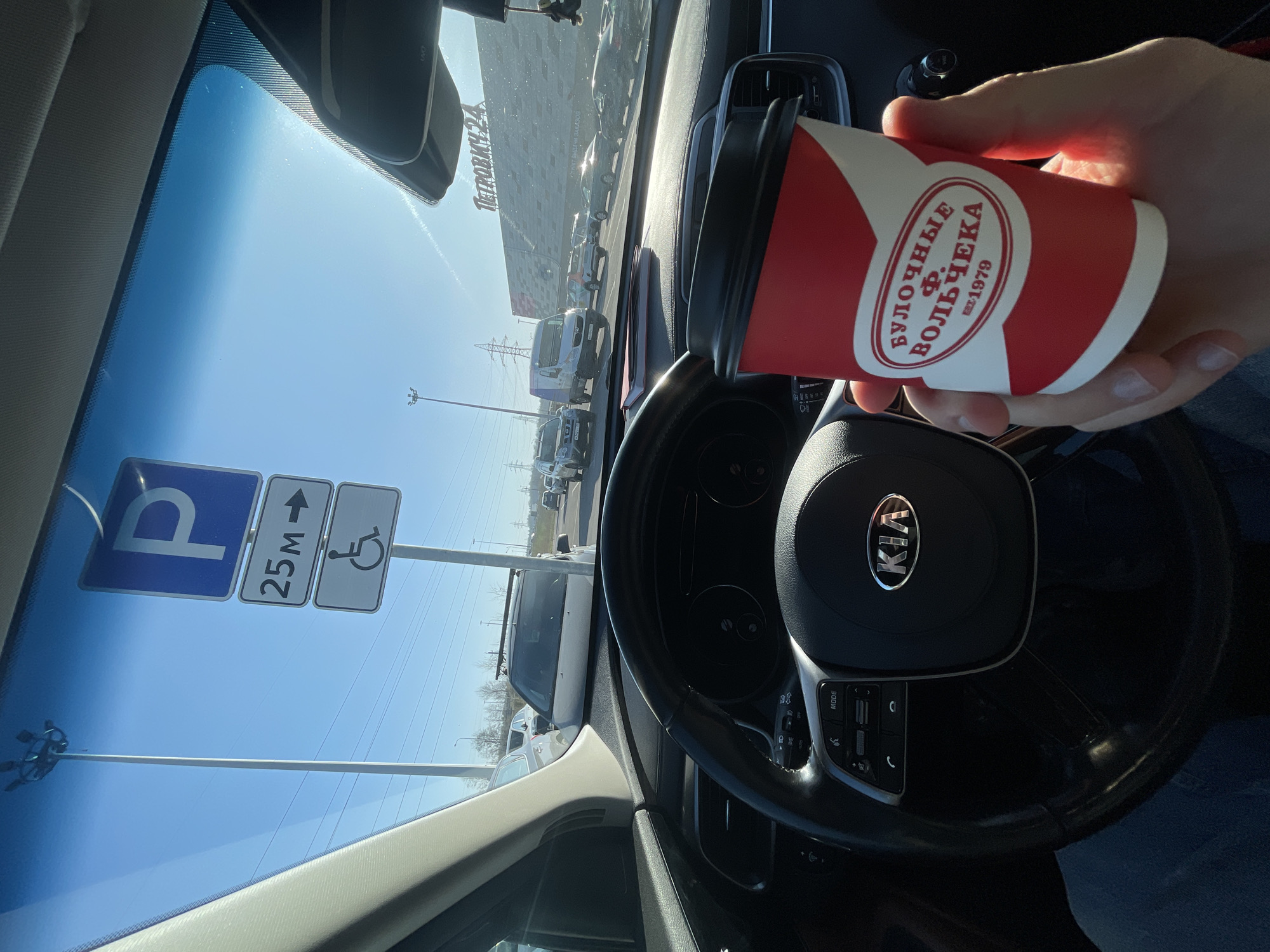 The image shows an interior view from a car, likely a Kia given the steering wheel logo. A hand is holding a paper coffee cup with the logo of "Булочные Ф. Вольчека" (Buloshnye F. Volcheka), an established bakery chain (est. 1979) from Saint Petersburg, Russia. Through the windshield, one can see a parking sign indicating parking for disabled individuals is 25 meters away, and parked cars. The reflection in the windshield suggests a large commercial building in the background. Given the presence of "Булочные Ф. Вольчека," the parking area sign, the parked cars and the context of Kudrovo, Russia, the location is possibly near the Mega Dybenko shopping center which houses a branch of "Булочные Ф. Вольчека" and is in the vicinity of Kudrovo. There is no specific person interaction. The action is holding the coffee cup while inside the car. The environment is a sunny day in a parking area in Russia.