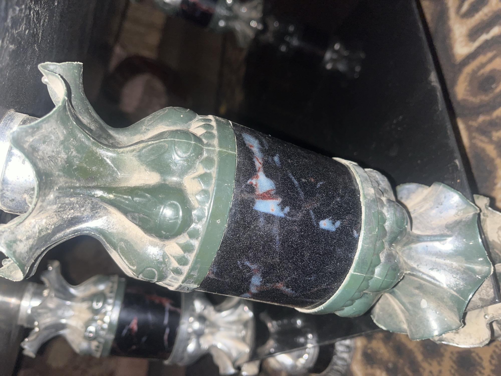 A close-up, slightly angled shot shows a dusty, decorative column with a dark, speckled cylinder in the middle. The column is made of a metallic material, likely silver-colored, and appears to be part of a larger piece of furniture or decor. It's coated in a layer of dust, particularly visible on the green, decorative elements and the silver-toned base and top. The central cylinder features a black, speckled surface with irregular patches of reddish-blue and white coloration, possibly representing a polished stone or faux material. The background is dark and out of focus, with hints of a reflective surface and a patterned rug. The scene is static and shows no people or activity. The objects are in a state of disuse or neglect, evidenced by the dust accumulation. The location is identified as Jalingo, Nigeria.