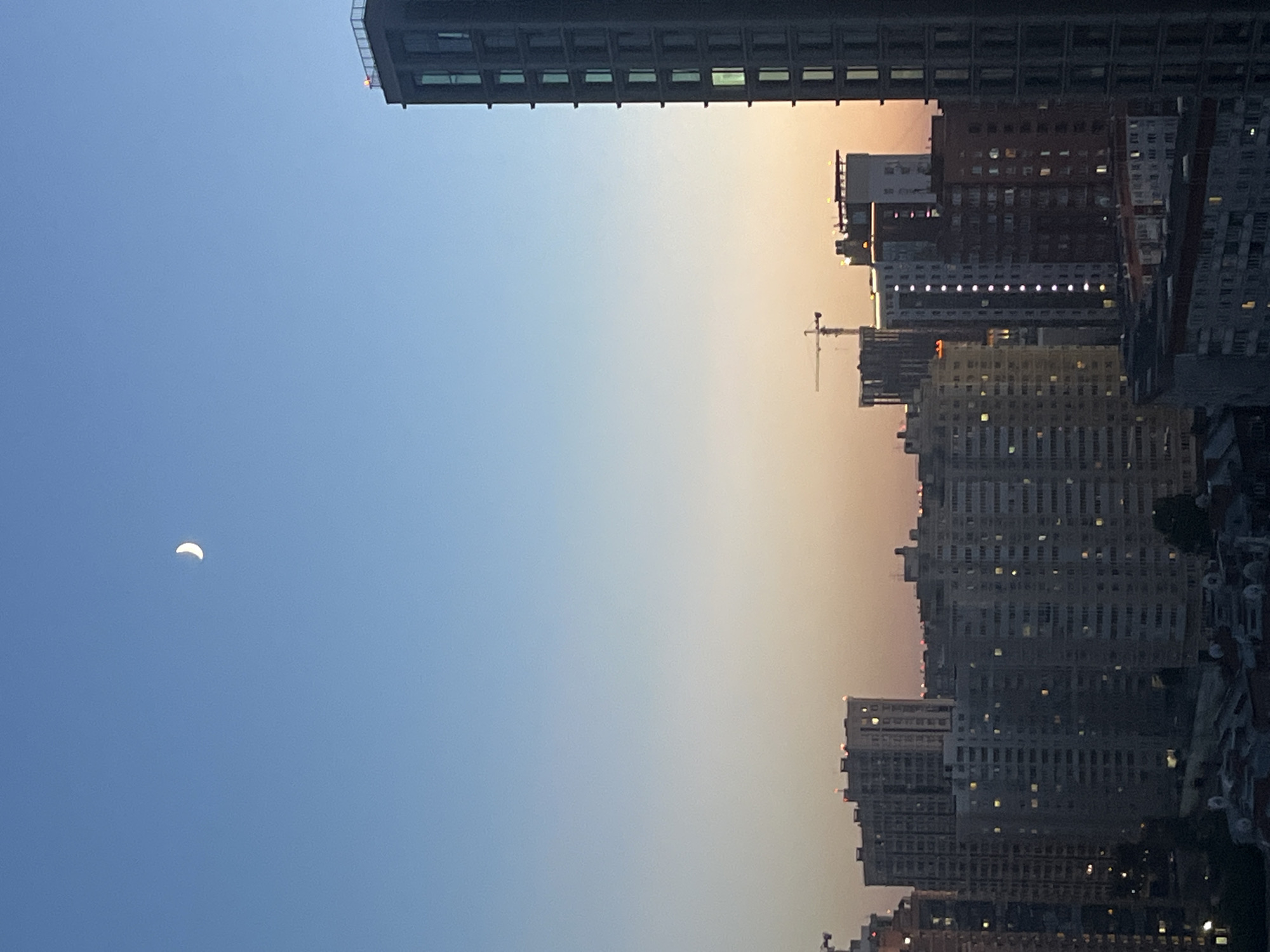 A crescent moon hangs in the clear blue sky above a city skyline at dusk. The sky transitions from a pale blue at the top to a soft orange and pink hue near the horizon, indicating the setting or rising sun. A cluster of tall, modern apartment buildings dominates the foreground and midground. The buildings vary in height and architectural style, with some featuring a more uniform, block-like design and others displaying a more contemporary, multi-faceted appearance. Many windows on the buildings are illuminated, suggesting occupancy. A construction crane is visible among the buildings. The image captures a sense of urban development against the backdrop of a twilight sky.