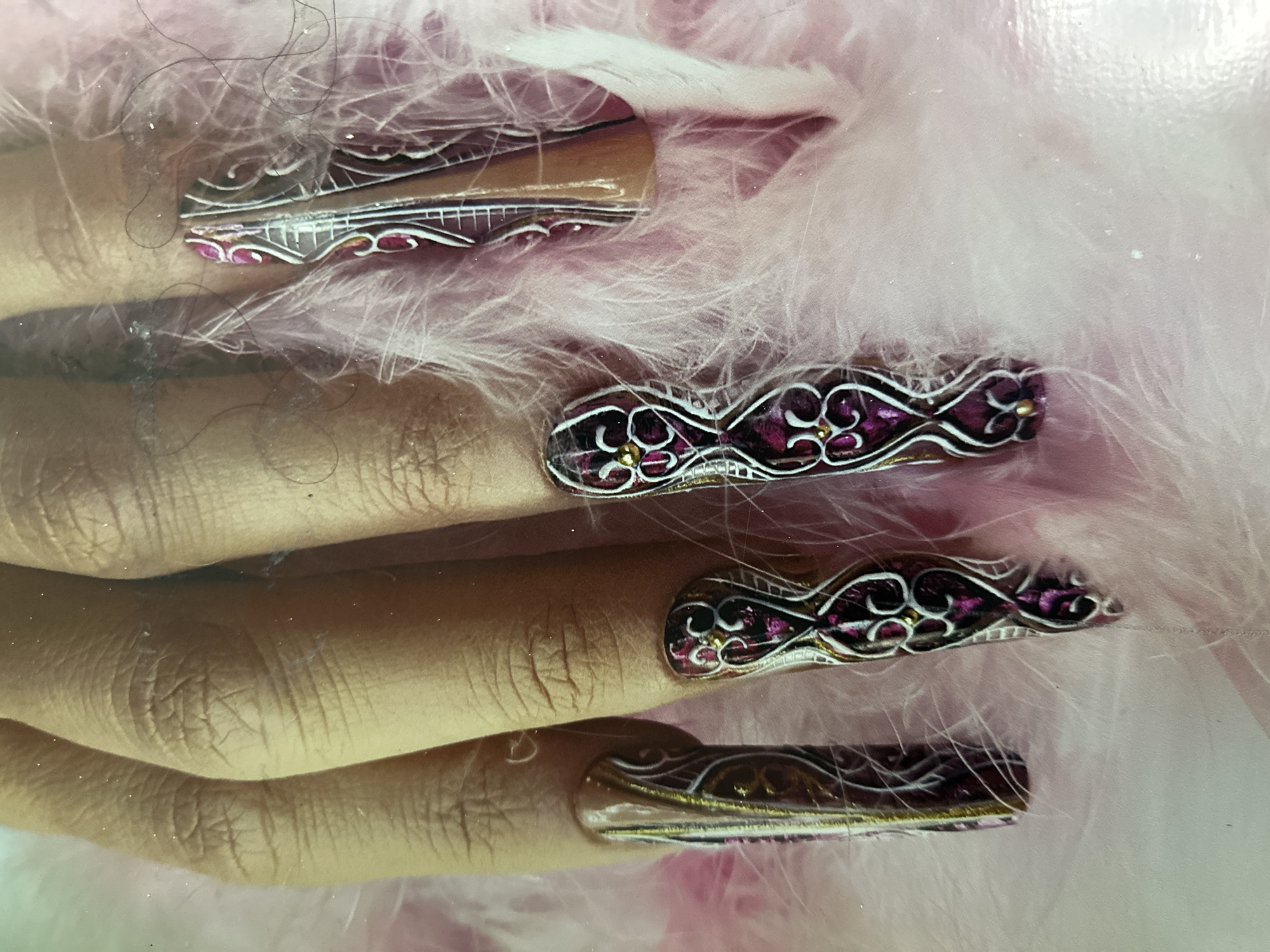 The image displays a close-up view of four elaborately designed artificial fingernails on a person's hand, which is partially visible. The fingernails are long and feature intricate patterns in shades of white, gold, and dark pink or purple. The patterns appear to be floral or scroll-like designs. The hand is resting on a fluffy pink material, possibly feathers or faux fur. There are some dark strands, likely hairs, scattered across the image, particularly around the fingernails. The lighting in the image is soft, highlighting the texture and detail of the nails and the surrounding material. The image does not provide any information about the location in Moscow, Russia.