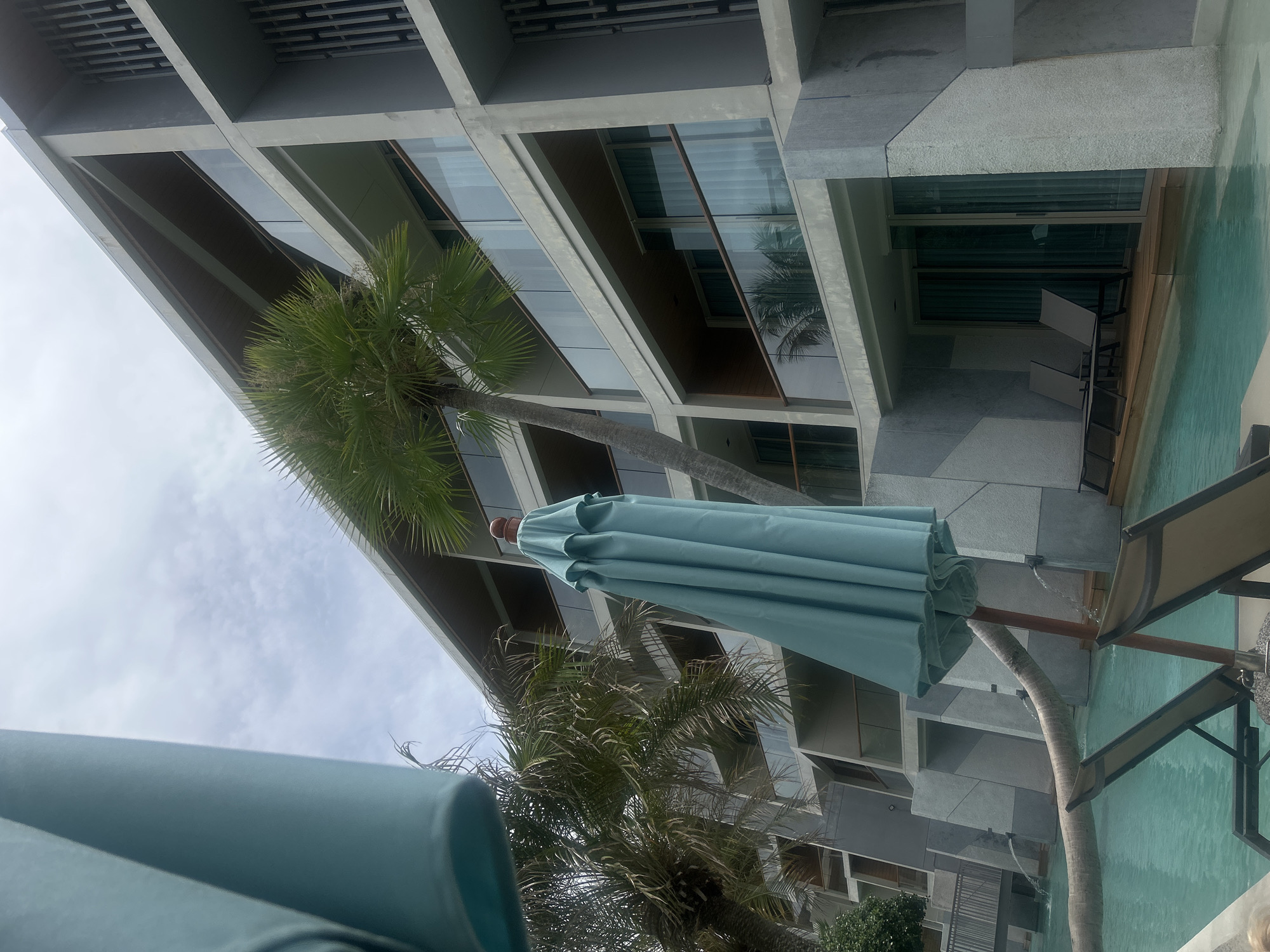 The image shows a view from a swimming pool in Rawai, Thailand. The pool water is light blue. A closed light blue umbrella is prominent in the foreground. Behind the umbrella, there is a building with a modern design, featuring multiple floors and recessed balconies. Palm trees are interspersed throughout the view, with some obscuring parts of the building. The sky is cloudy, with some patches of blue visible. There are lounge chairs by the pool.