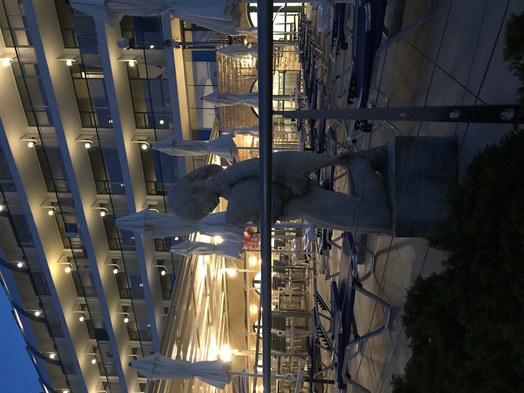 The image depicts an outdoor terrace area of a hotel or resort, likely in Varna, Bulgaria. In the foreground, a stone sculpture of a person is visible, partially obscuring the view of rows of empty blue lounge chairs and closed white umbrellas. Behind the terrace, a multi-story building with balconies and illuminated windows is seen against a twilight sky. The building's architecture suggests it is a hotel, possibly with a resort-style ambiance. The terrace itself is paved with tiles, and a metal railing runs across the middle ground, separating the lounge area from what appears to be a walkway or dining area. The lighting suggests it is either early morning or late evening. No people are clearly visible in the scene, but the arrangement of furniture implies it is a place designed for leisure and relaxation. The overall impression is one of a quiet, possibly off-season, resort setting.