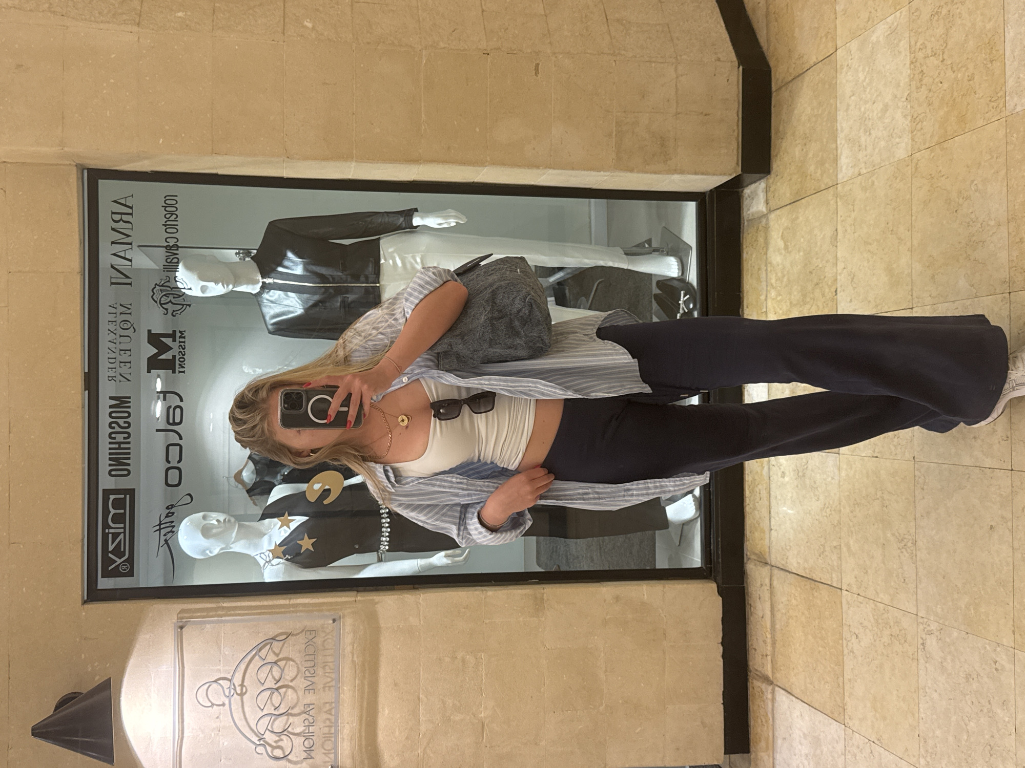 A young woman is taking a selfie in front of a display window. She is wearing navy blue pants, a white crop top, and a light blue striped button-down shirt. Sunglasses are tucked into the top of her shirt. She is holding a phone up to her face in her right hand.
The display window behind her features mannequins dressed in designer clothing. Logos for "Moschino" and "Armani" are visible on the window.
The location appears to be a shopping mall or high-end retail area. The floor is tiled. A sign with "Exclusive Fashion" in reversed text is visible below the window. There is no information available in the image to confirm or deny its location in Herzliya, Israel.