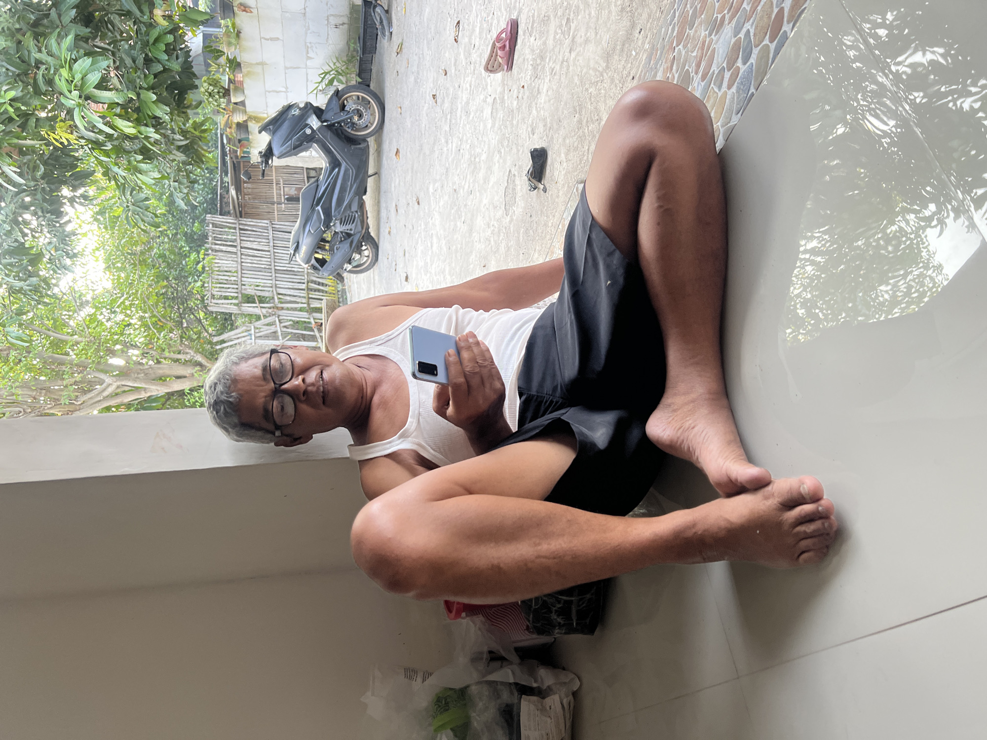 An elderly man with graying hair and glasses is sitting on the ground in front of a building, holding a smartphone. He is wearing a white tank top and black shorts. The background shows a courtyard with a motorcycle parked, some potted plants, and a bamboo fence. The scene appears to be in a residential area in Kecamatan Pati, Indonesia, suggested by the architecture and vegetation.