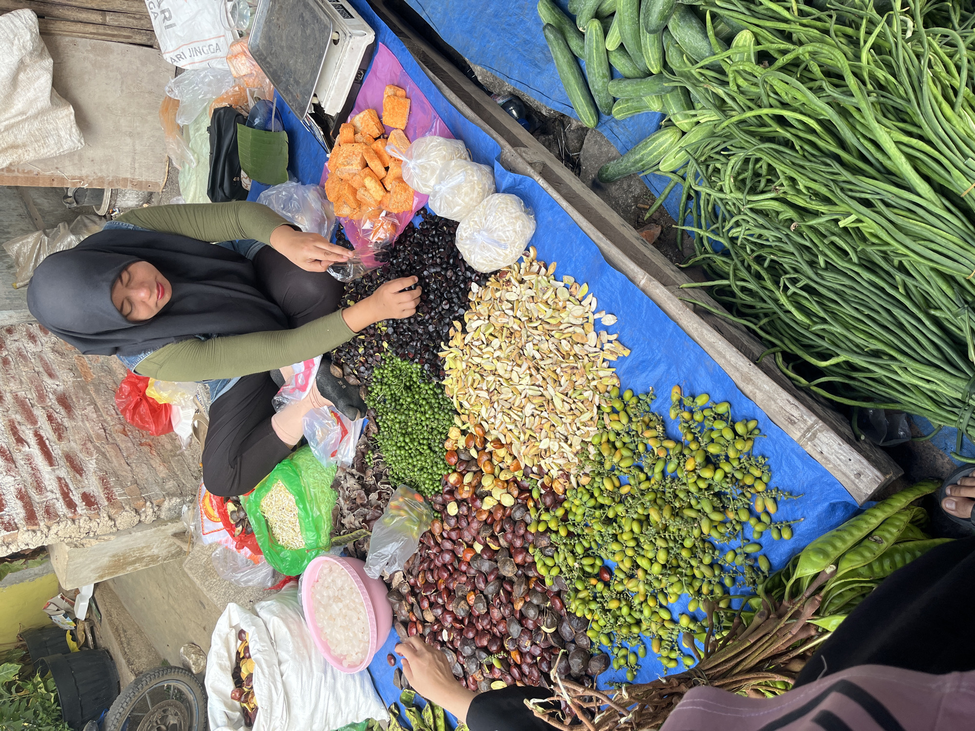 A vendor, wearing a black hijab and green shirt, kneels beside a blue tarp spread on the ground in what appears to be an outdoor market in Kemuning, Indonesia. The tarp displays a variety of fresh produce, including piles of green beans, cucumbers, various seeds and nuts, and some orange-colored food items wrapped in plastic. The vendor is actively engaged in her work, as indicated by her hands sorting through produce and a weighing scale positioned nearby. The backdrop reveals a brick wall and some potted plants, suggesting a bustling urban marketplace environment.