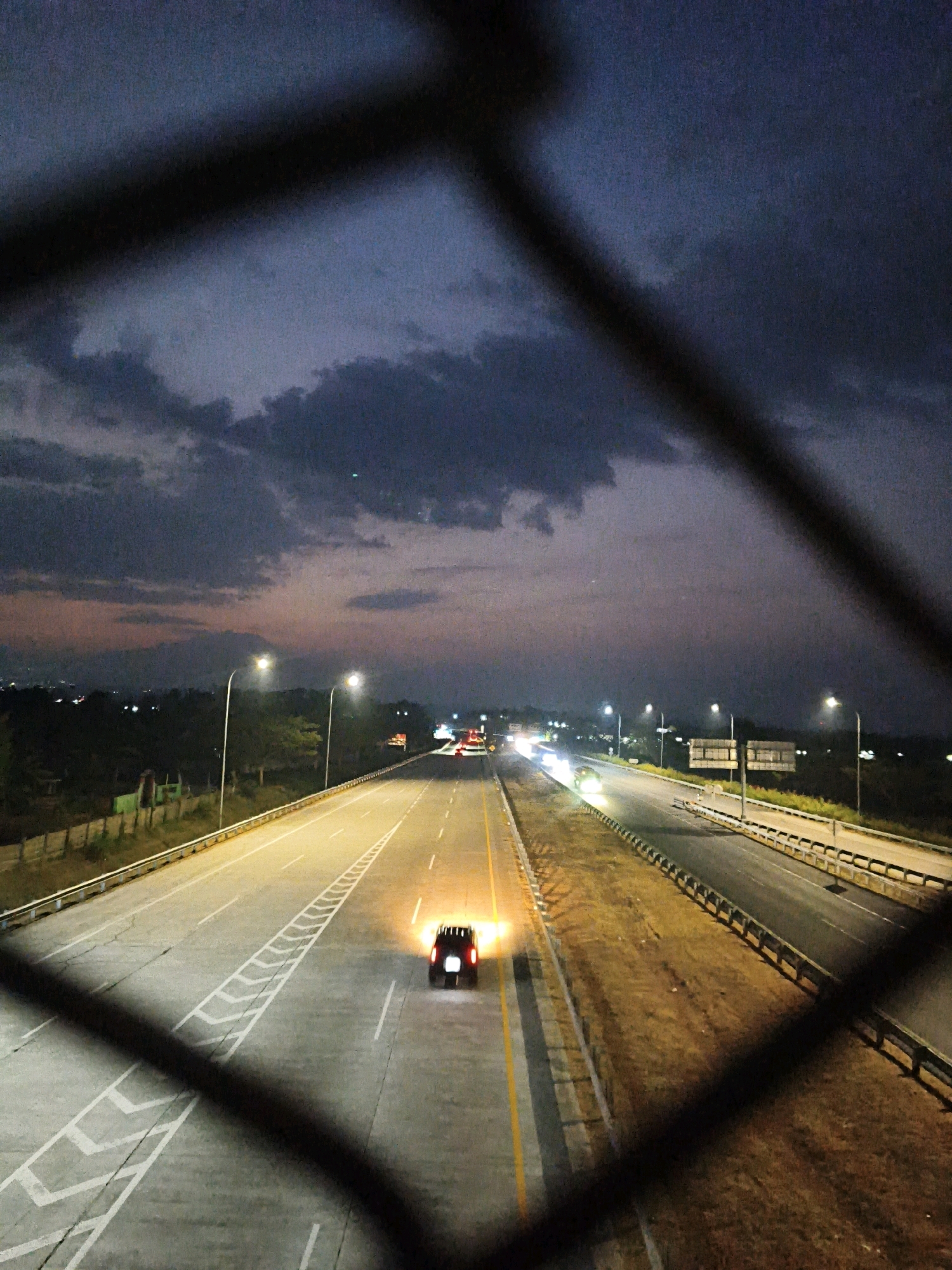The image depicts a highway at dusk, likely the Ngargorejo toll road in Indonesia, viewed through a wire fence. The sky is a dark, cloudy blue with hints of purple and orange near the horizon. The highway is illuminated by streetlights, casting a warm glow on the asphalt. A dark-colored car, possibly an SUV, is traveling on the highway with its headlights and taillights illuminated, creating bright flares of light. Other vehicles and distant lights are visible further down the road, suggesting ongoing traffic. The landscape on either side of the highway is lined with trees and vegetation, with some structures visible in the distance. The fence in the foreground, made of dark metal wires, partially obstructs the view, creating a layered effect.