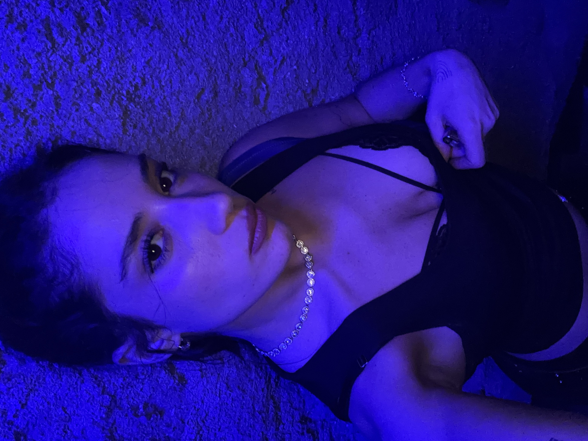 A young adult woman lies on a textured surface under intense blue lighting. She wears a black top with lace trim and a delicate silver necklace. A bracelet is visible on her wrist. Her gaze is directed toward the viewer, and she appears to be adjusting the neckline of her top with her hand. The background is undifferentiated due to the lighting and close-up composition, preventing location identification within Buenos Aires or any other specific place.