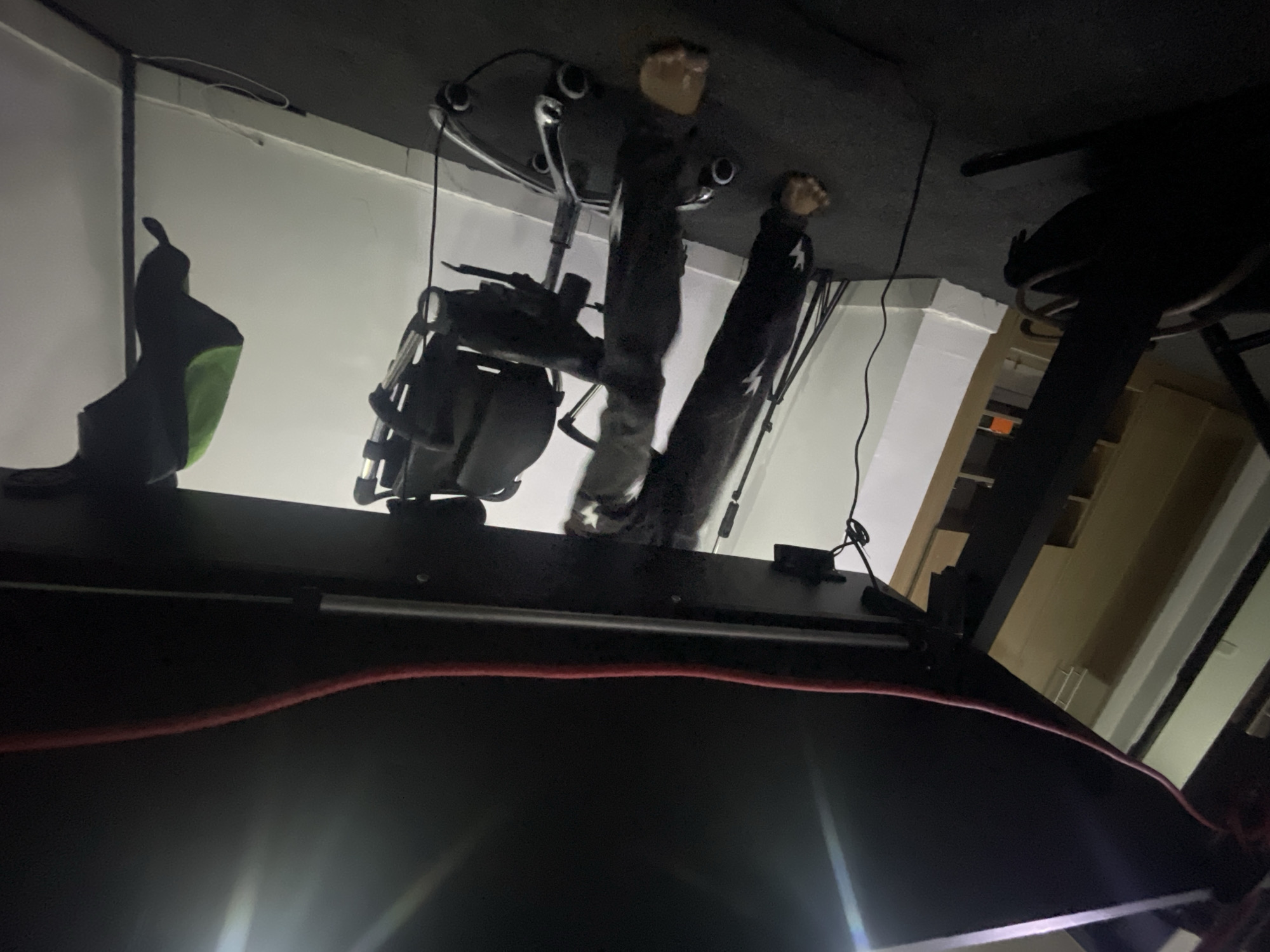 The image is taken from under a desk or table in an indoor environment. Above, we see a dark office chair with two human figurines attached to the bottom. The figurines appear to be wearing dark clothing with white stars on them. One is standing upright, while the other is leaning. The chair is attached to a base with multiple legs. To the left of the chair is a green and black bag hanging from the desk edge, as well as a black object. The desk itself is black, with a red cable running across it. The environment is dimly lit, with bright light reflecting off the desk surface. In the background, there is a white wall and the suggestion of an open doorway or a window. The location is not determinable from the image.