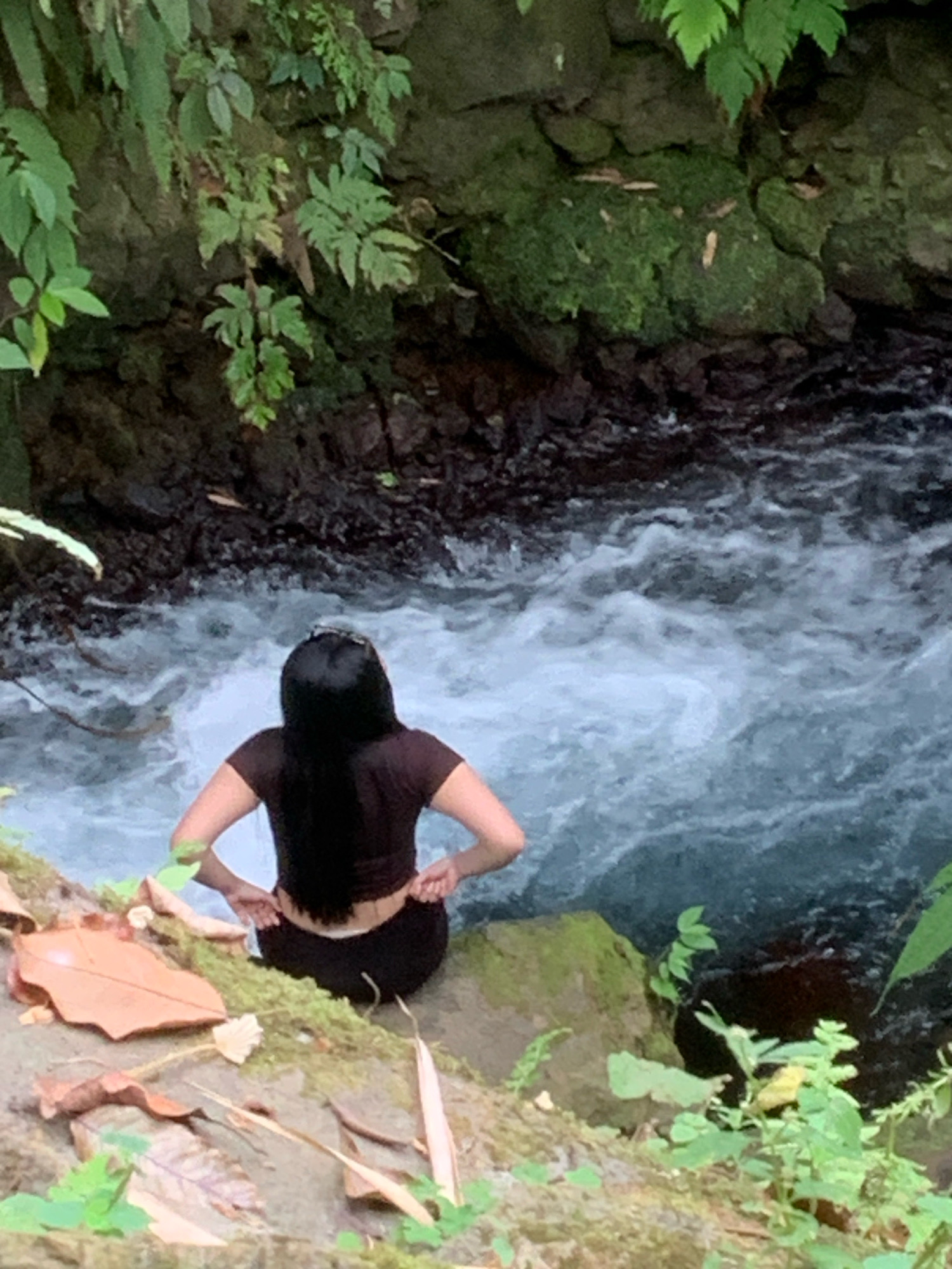 A person with long black hair, wearing a dark brown t-shirt and black pants, is sitting on the edge of a river in Pronojiwo, Indonesia. They are facing away from the camera, looking out at the fast-flowing water. The riverbed is rocky and dark, with turbulent white water indicating a strong current. The river banks are lined with lush green vegetation, moss-covered rocks, and fallen leaves. In the background, dark rocks and dense foliage form a natural backdrop. The scene appears to be in a natural, wild environment.