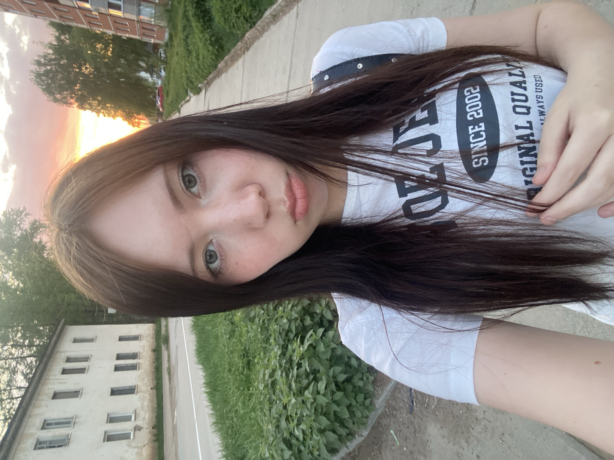 A medium-angle self-portrait of a young woman with long, dark brown hair that is partially obscuring her face. She has fair skin, light-colored eyes, and visible freckles. She is wearing a white T-shirt with black lettering that reads "SINCE 2002 ORIGINAL QUALITY ALWAYS USED." The background features urban elements, including a building with multiple windows, a concrete sidewalk, and greenery. The lighting suggests a sunset or early evening, with a warm glow visible in the upper left corner. The location appears to be an outdoor space near residential buildings. The city of Unknown, Unknown is unknown.