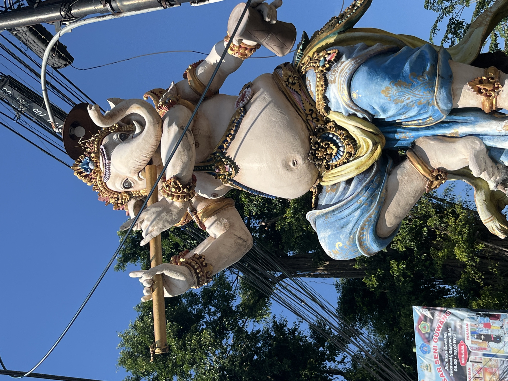 This image captures a vibrant, larger-than-life statue of Ganesha, a prominent deity in Hinduism, situated outdoors against a bright blue sky and lush green trees. The statue is richly adorned with intricate golden jewelry and colorful clothing, featuring a blue dhoti with ornate gold patterns. Ganesha is depicted playing a flute, with one hand holding the instrument and the other near his mouth. In his right hand, he holds a small drum. The statue appears to be part of a larger display or event, possibly a parade or festival, given the festive decorations.
In the lower left corner, a banner is visible with text indicating "SAR SENI GUWANG," which translates to "Guwang Art Market." Below this, an address is printed: "Jln. Raya Guwang, Sukawati, Gianyar, Bali." This clearly places the scene in Sukawati, Gianyar, Bali, Indonesia. The banner also lists operating hours: "08.00 - 18.00 Wita" and advertises "MENJUAL" (selling) "PAKAIAN" (clothing) and "KERISAN" (likely referring to traditional Balinese crafts or arts, such as keris daggers, though the spelling might be slightly varied).
Overhead, a network of electrical wires and a utility pole are visible, suggesting an urban or semi-urban setting within Sukawati. The overall impression is one of cultural richness and artistic expression, typical of Bali.