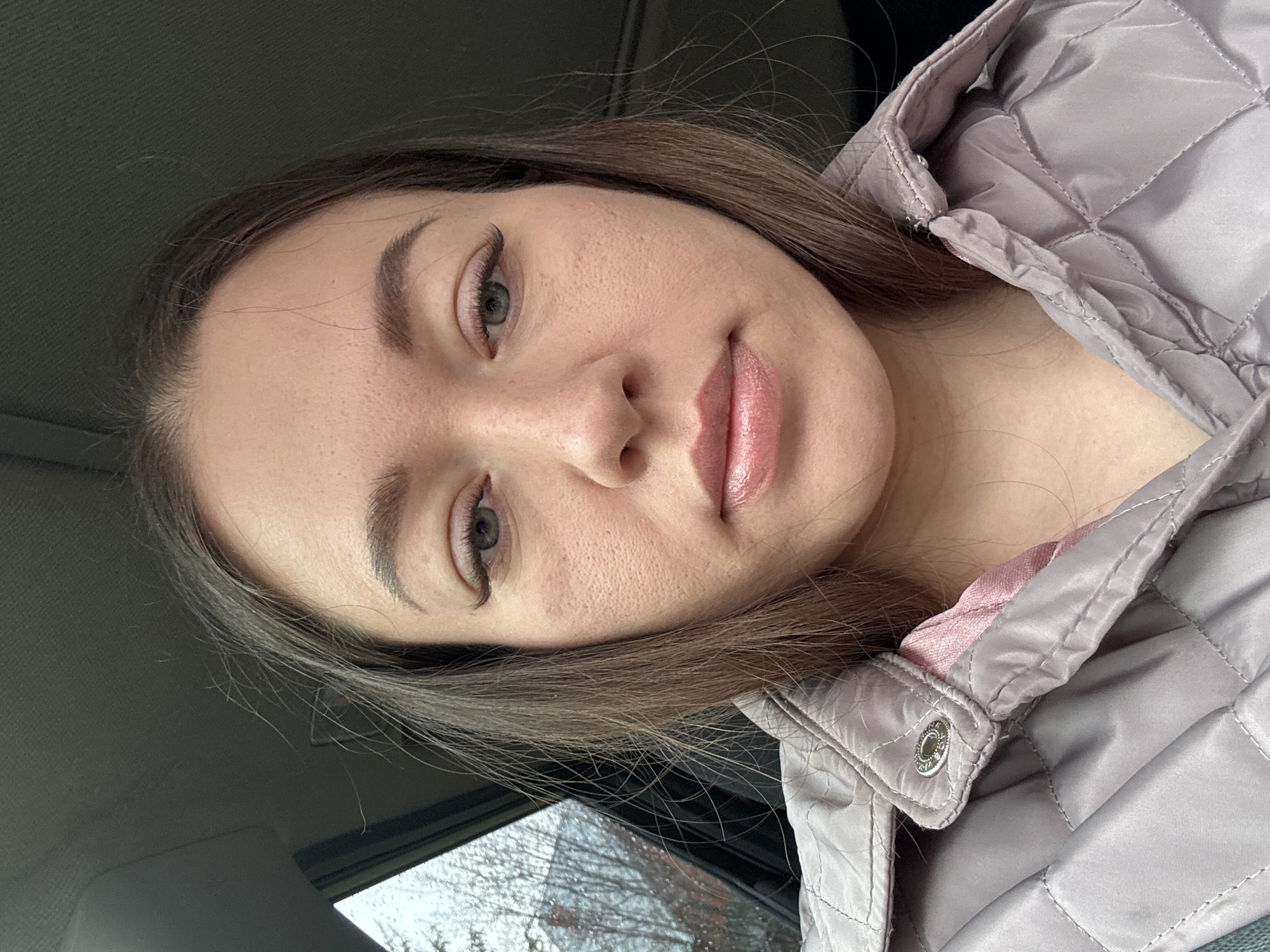 The image shows a woman's face from a close, angled perspective. She has light skin, brown hair, defined eyebrows, and green eyes. She is wearing a light purple quilted jacket with pink lining. The background appears to be the interior of a car. It is not possible to confirm the location in the city of Ферма, Russia from the given image.