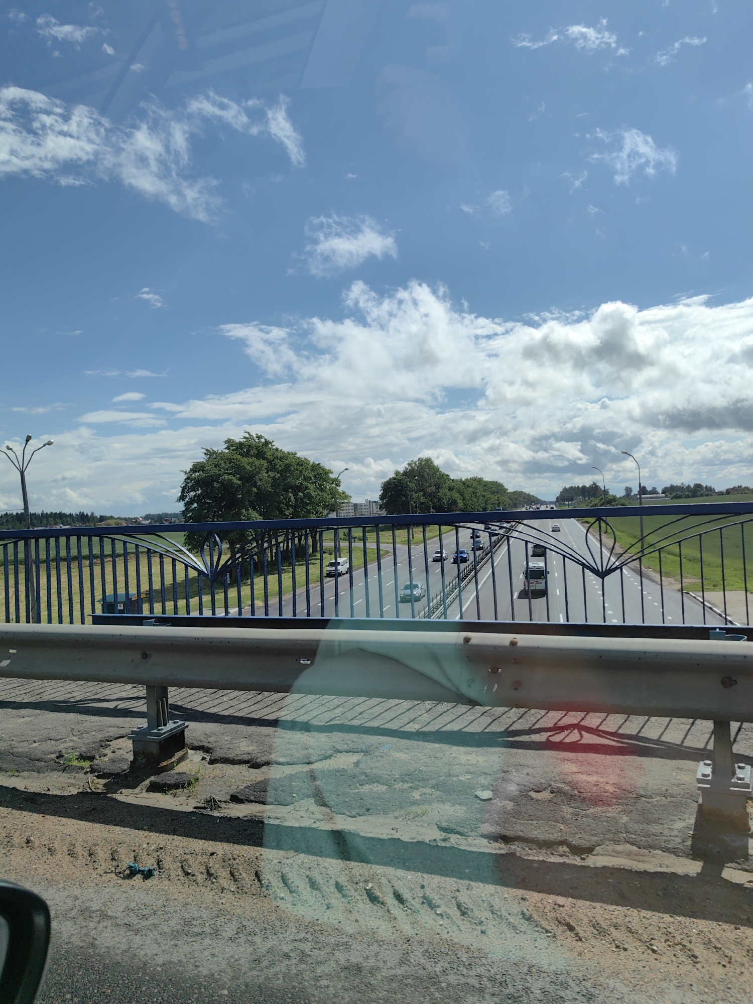 The image captures a view from an elevated position, likely a bridge, overlooking a highway in the vicinity of Фаніпальскі сельскі Савет, Belarus. The sky is blue with scattered white clouds. Below, a multi-lane highway stretches into the distance, with several cars and a white van in motion. On either side of the highway, there are green fields and clusters of trees. A blue metal fence runs along the edge of the elevated viewpoint, obscuring some of the foreground. The ground in the immediate foreground appears to be a rough, uneven surface, possibly asphalt with some damage. The lighting suggests it is daytime, likely midday given the brightness and shadows.