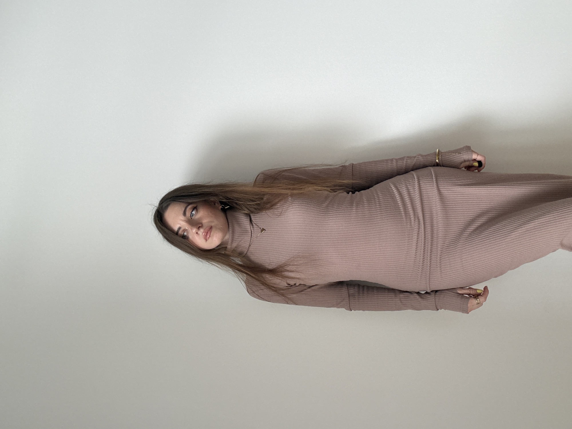 A woman is seen in a full-length shot wearing a long-sleeved, ribbed turtleneck dress of a muted mauve color. She has long, straight brown hair and is accessorized with gold jewelry including a necklace and bracelet. Her pose is slightly angled, with her head toward the left side of the frame. The background is a plain, light-colored wall. There are no indicators to suggest this image was taken in Moscow, Russia.