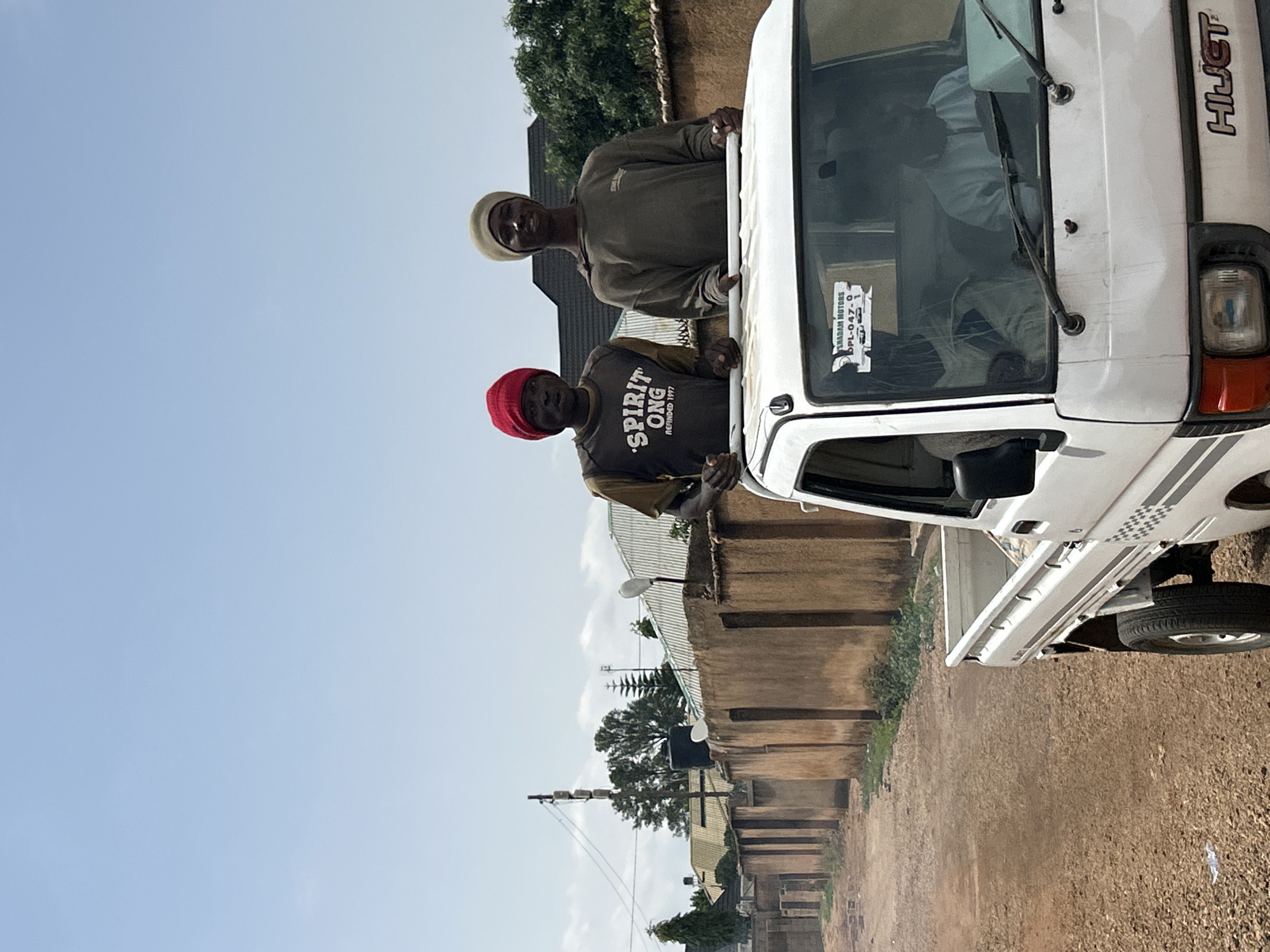 Two men are standing in a white utility truck with the "HIJET" logo visible. One man wears a red hat and a dark t-shirt printed with "Spirit ONG." The other man wears a light-colored hat and a long-sleeved shirt. The location appears to be a street in Jos, Nigeria. The background shows residential buildings, foliage, and power lines. The truck is parked on a dirt road.