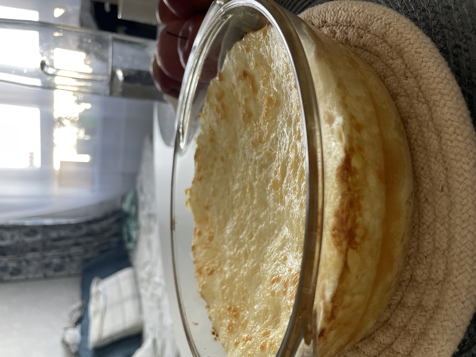 A tilted, close-up shot shows a glass baking dish filled with food, being held with a beige woven hot pad or trivet. The food appears to be a baked dish with a cheesy or bubbly topping. Part of a hand and some red objects (possibly apples) are visible at the top of the image. The background is blurry and shows a room with some furniture and a window. There is no evidence in the picture to suggest the location is Izhevsk, Russia.