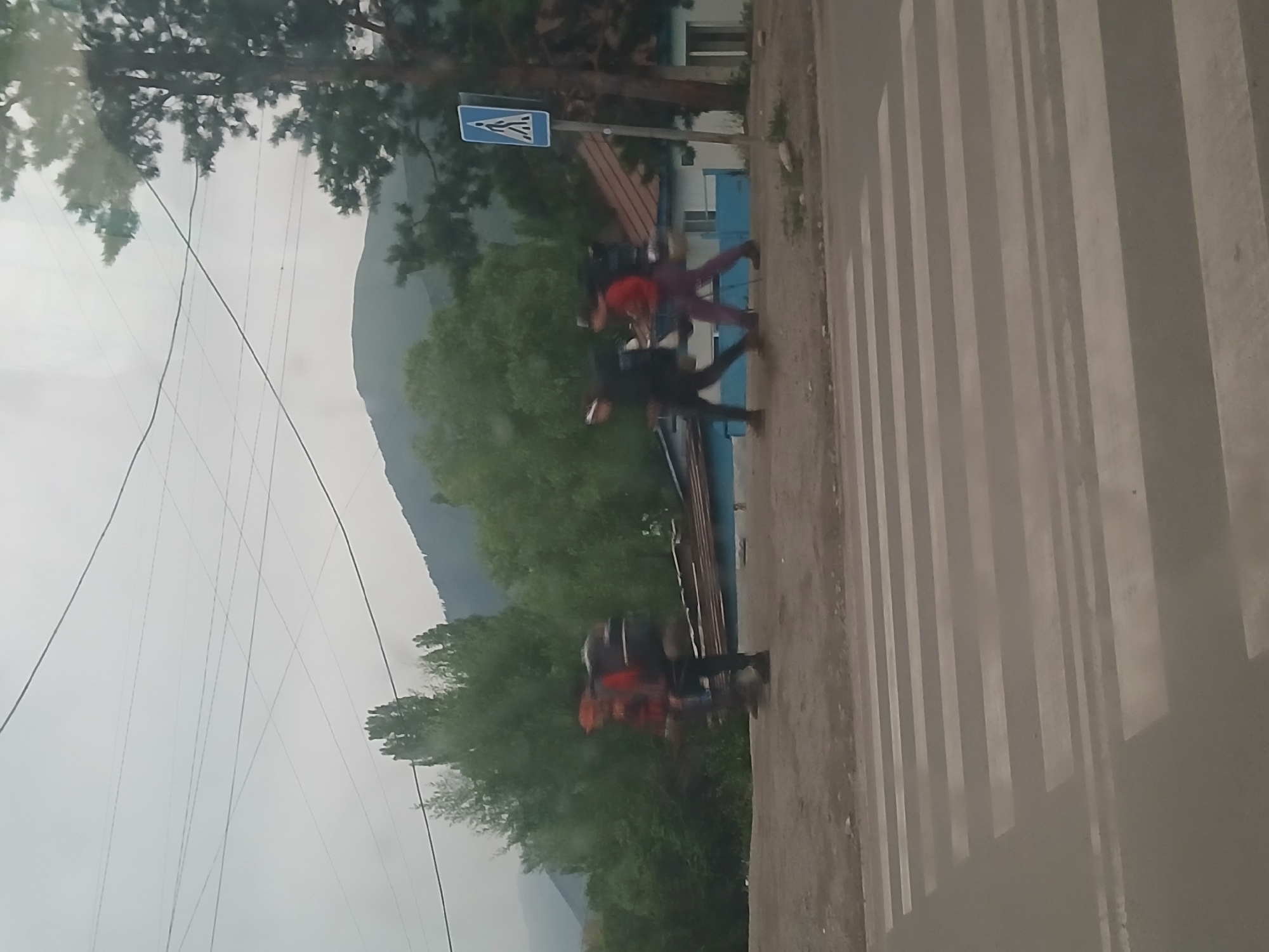 The image shows a pedestrian crossing in a semi-urban or rural area, with a overcast sky. In the background, there are trees and mountains. Three individuals are walking across the pedestrian crossing, all wearing backpacks, suggesting they are hikers or travelers. The person on the left appears to be wearing a red shirt and dark pants. The person in the middle is wearing a dark top and dark pants. The person on the right is wearing a red top and dark purple pants. A pedestrian crossing sign is visible to the right of the scene. The overall impression is of a cloudy day in a mountainous region. It is plausible that this scene is located in Ak-Suu, Kyrgyzstan, given the natural landscape.