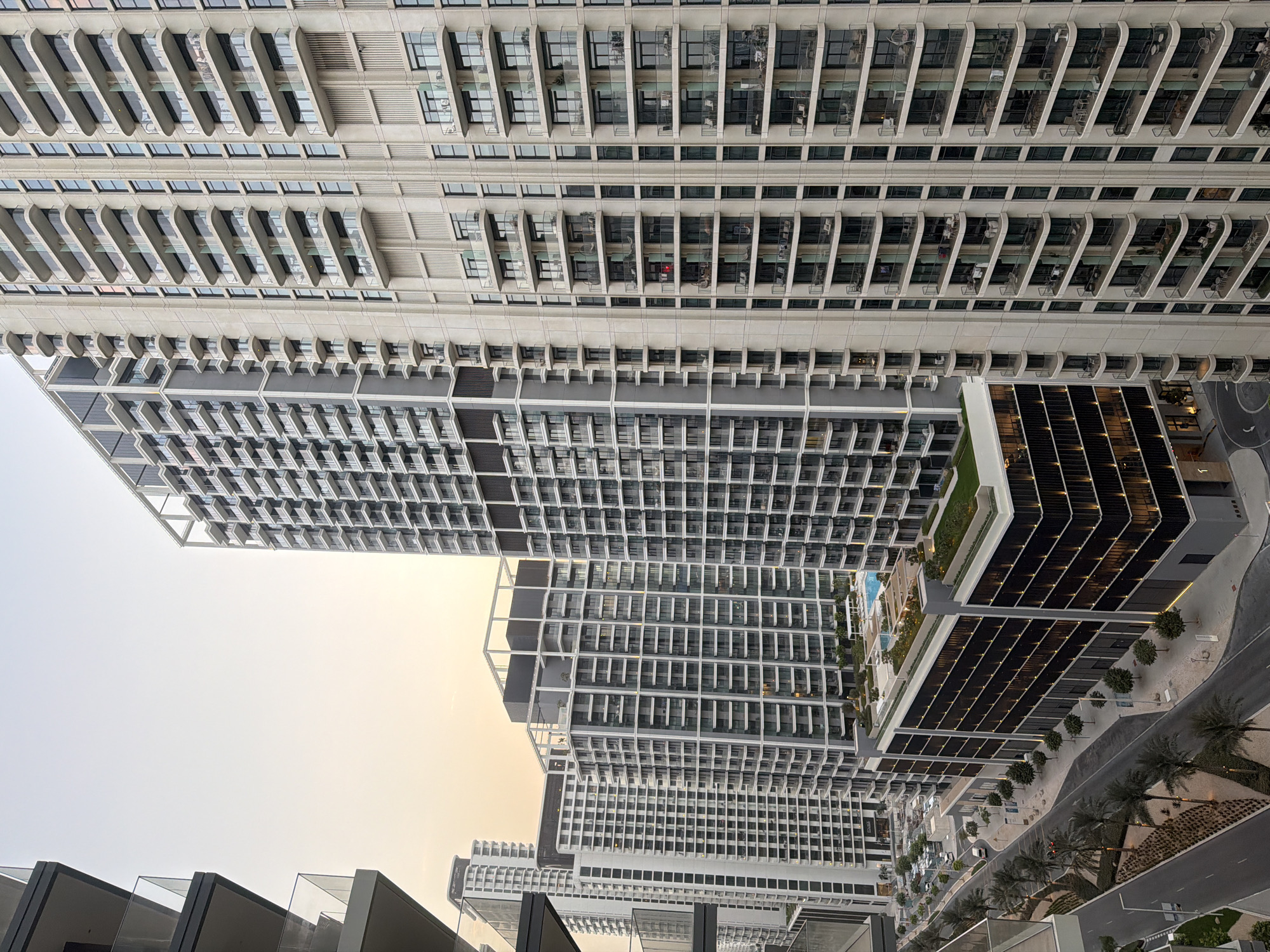This is a daytime, eye-level, medium shot of a modern urban cityscape in Dubai, United Arab Emirates, with a focus on the architecture of high-rise residential buildings. The buildings are predominantly light-colored with repeating patterns of balconies and windows. Several buildings are visible, including a very tall one on the right with a light beige facade and numerous balconies. To its left is another tall building with a darker gray facade. In the foreground, a lower building with a dark facade is visible, featuring rows of illuminated windows. A swimming pool with blue water is situated on the roof of this lower building, surrounded by greenery. A street with palm trees and landscaped areas runs along the left side of the image, with a few cars and streetlights visible. The sky is a pale, overcast orange, suggesting either dawn or dusk. There are no people clearly visible in the image.