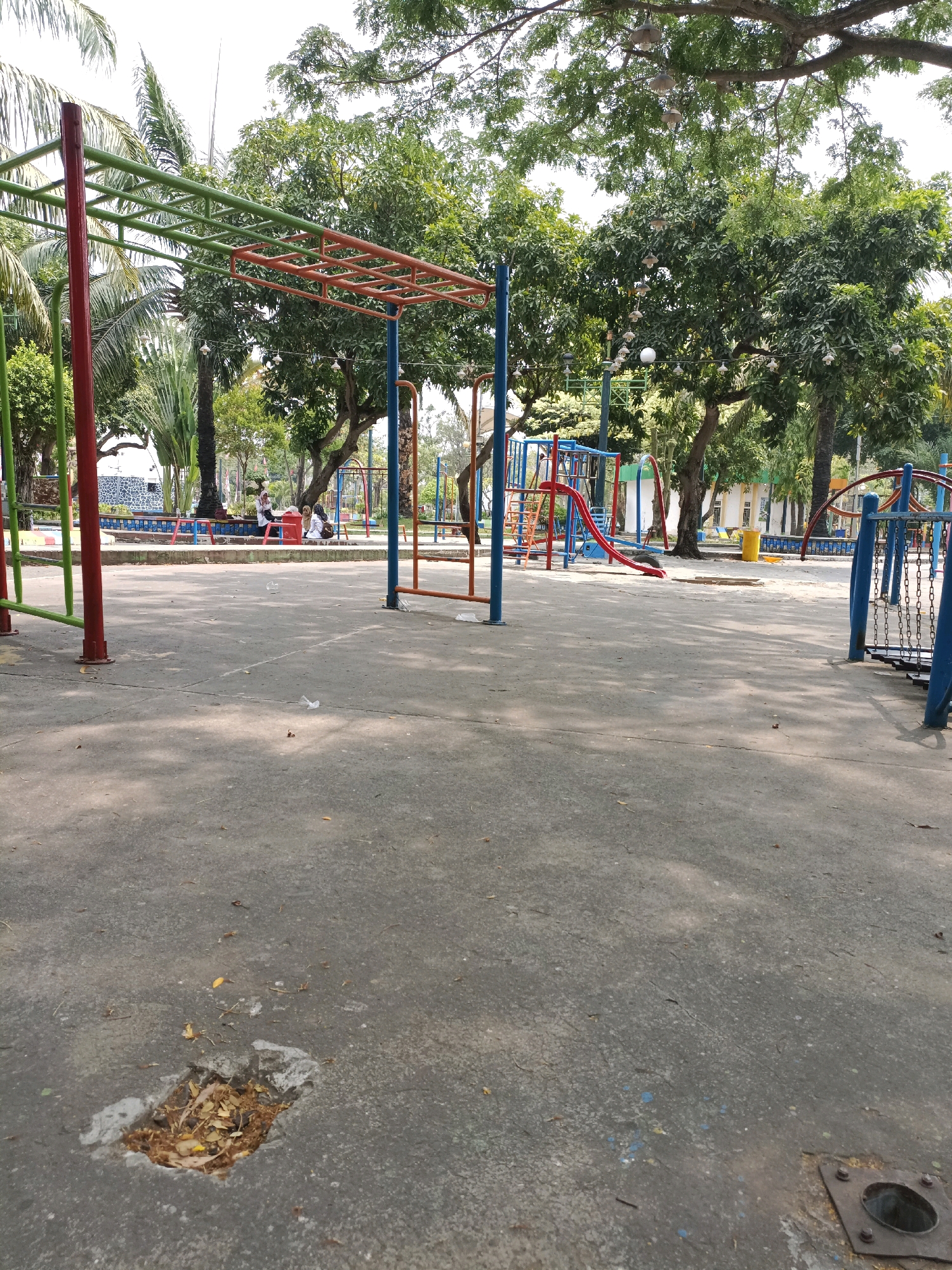 The image depicts a deserted outdoor playground with a concrete ground and scattered leaves. In the background, there is a slide, monkey bars, and other playground equipment. Lush green trees and palm trees surround the playground, with some string lights hanging from the branches. There are a few people sitting on a bench in the distance. The lighting suggests it is daytime. The image appears to be taken from a low angle, looking across the playground. There are some minor imperfections on the concrete, including a small hole filled with leaves and debris, and a metal fixture on the ground in the foreground. The overall scene is one of a quiet, perhaps underutilized, public space.