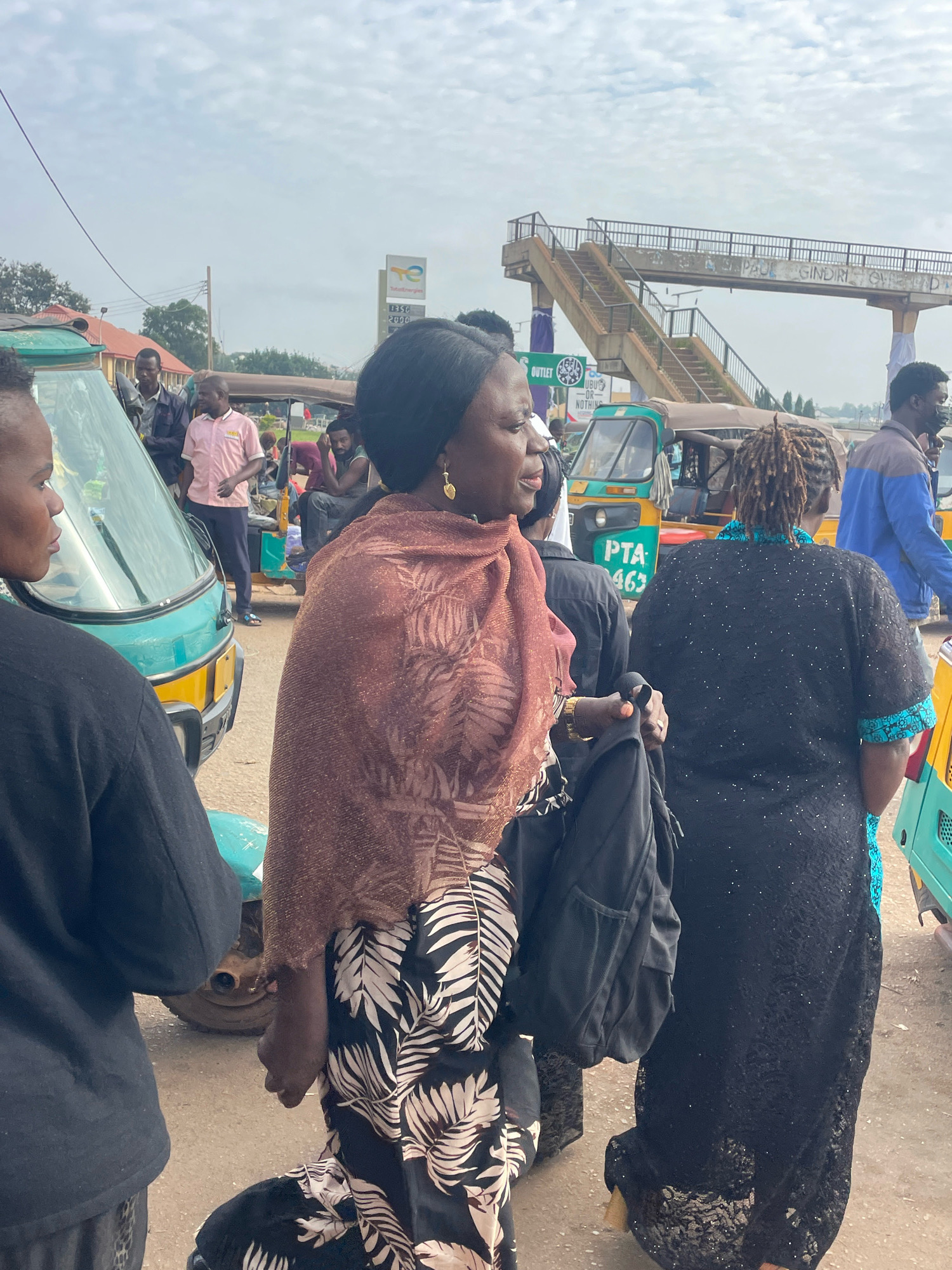 In Gura Top, Nigeria, a group of individuals are seen near a road intersection. A woman in the foreground, wearing a brown patterned shawl over a black and white floral dress, walks with a black backpack slung over her shoulder. Beside her, a woman in a black lace dress with blue accents is visible. To their left, another woman in a black top and patterned pants is partially obscured. In the background, several auto-rickshaws are present, with one prominently displaying "PTA 463". A pedestrian bridge spans the road overhead. Additional individuals are seen standing and sitting near the auto-rickshaws. The environment appears to be a busy, outdoor urban setting with a cloudy sky.