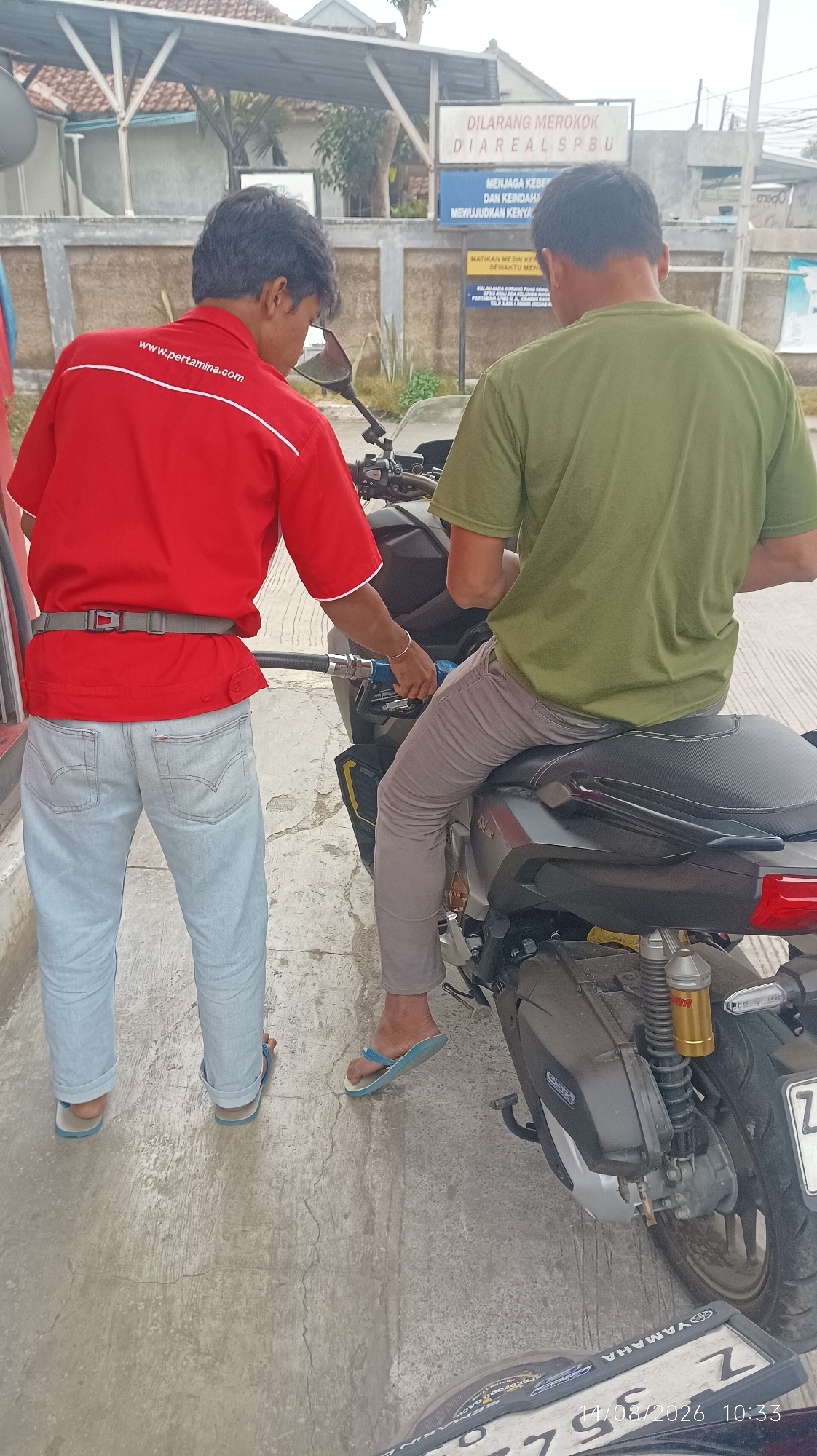 Two men are at a gas station in Tasikmalaya, Indonesia. One man, wearing a red shirt with "www.pertamina.com" printed on it and light-colored jeans, is standing and operating a gas pump. The other man, wearing a green t-shirt and gray pants, is sitting on a motorcycle. The motorcycle is black with some yellow accents. The ground is paved and appears wet in some areas. There is a sign in the background with Indonesian text. The license plate of the motorcycle is partially visible, showing the letter "Z". A date and time, "14/08/2026 10:33", is superimposed on the lower portion of the image, indicating it is likely a timestamp from a device.