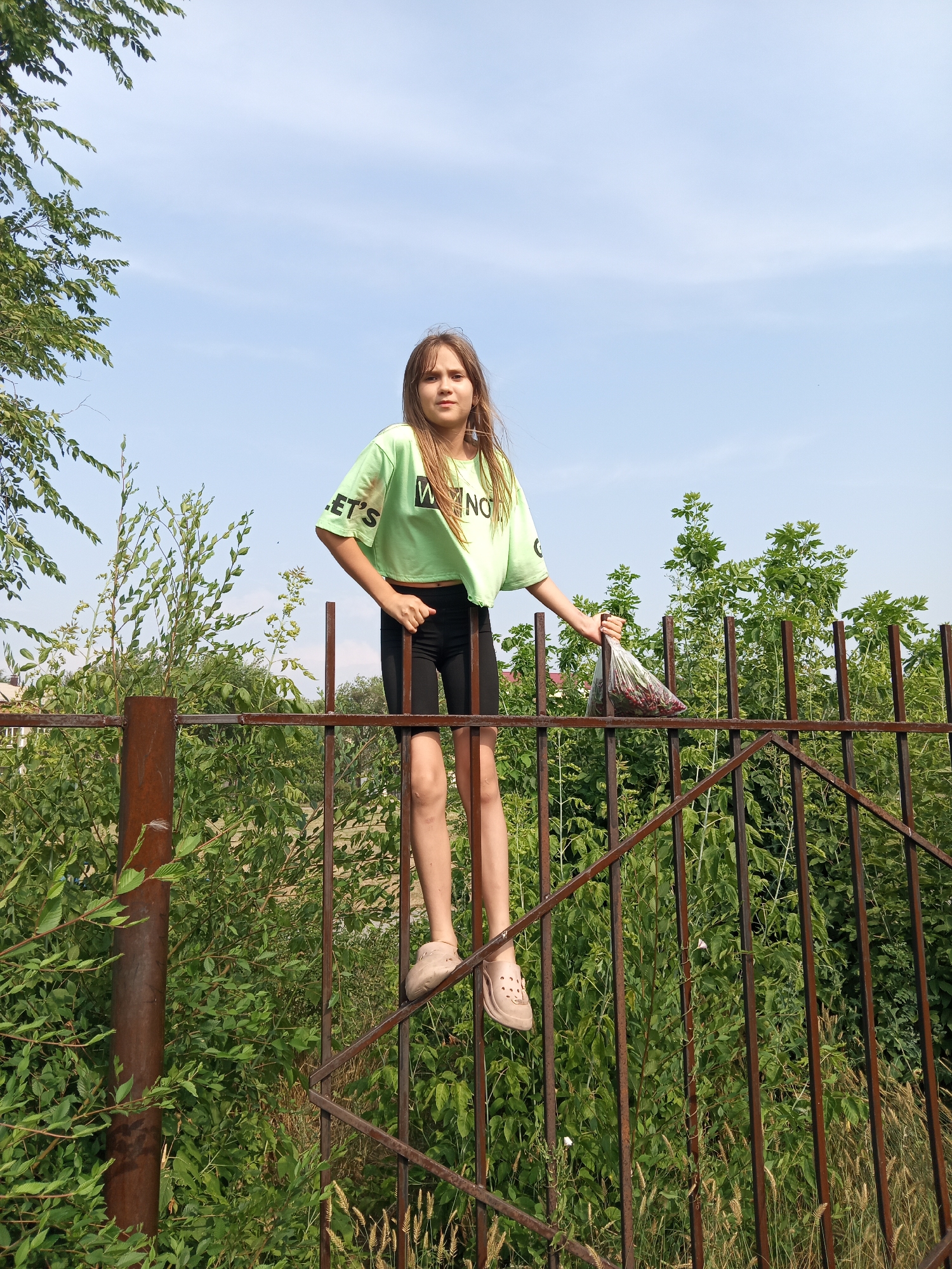 A young girl with long, light brown hair, wearing a lime green crop top with black lettering and black athletic shorts, stands on top of a rusty metal fence. She is also wearing light brown Crocs. The girl is holding a clear plastic bag filled with red berries in her right hand, which she extends forward. The fence is made of vertical metal bars and has a diagonal section. Behind the fence, there is dense green foliage and trees. The sky above is blue with scattered white clouds. The scene appears to be outdoors, likely in a residential area, given the presence of a fence and vegetation. There are no other individuals visible in the image. The girl's left hand is resting on the fence. The overall lighting suggests it is daytime. The girl is facing the camera, looking directly at it.