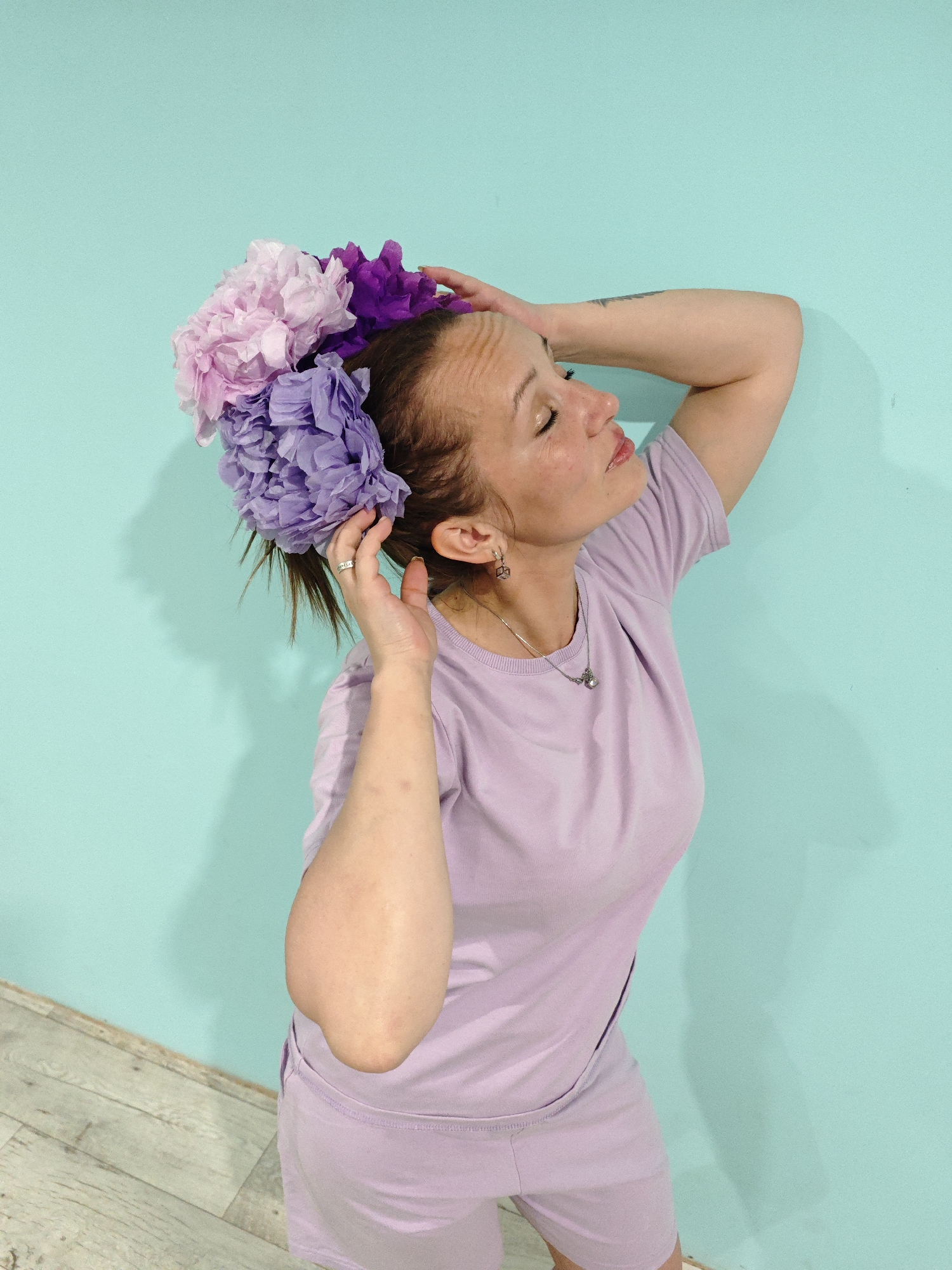 A full-length shot of a woman indoors standing against a bright turquoise wall with a light wood floor at the bottom left corner. She is looking up and to the right. She is wearing a light lavender matching T-shirt and short set. She has multiple pink and purple flower decorations in her hair. Her left arm is bent and her left hand is touching the flowers in her hair near her left ear. Her right arm is bent behind her head with her right hand near the back of her head. She has a necklace and earring set. Freckles are visible on her face.
There is no indication of this image being in the city of Markovo, Russia.