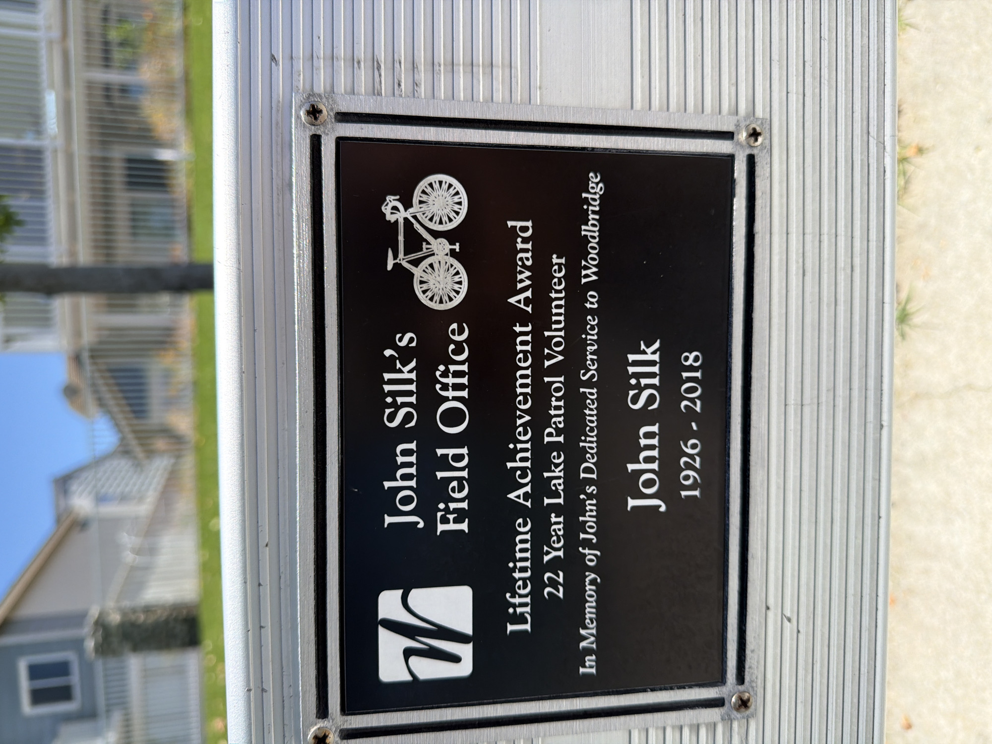 The image shows a close-up of a metal plaque mounted on a silver-colored case. The plaque is black with white text and graphics. At the top left of the plaque is a white square with a stylized "W" inside. To the right of the "W" is the text "John Silk's Field Office". Below this, centered on the plaque, is the text "Lifetime Achievement Award", followed by "22 Year Lake Patrol Volunteer", and then "In Memory of John's Dedicated Service to Woodbridge". The bottom of the plaque features the name "John Silk" in larger font, followed by the years "1926 - 2018". A white graphic of a bicycle is positioned to the right of the text "Field Office". The plaque is secured to the case with four screws, one in each corner. The case itself appears to be made of ribbed metal. The background of the image is out of focus and shows a grassy area, a white fence, and residential buildings, suggesting an outdoor suburban setting. The lighting suggests it is daytime. No people are visible in the image.