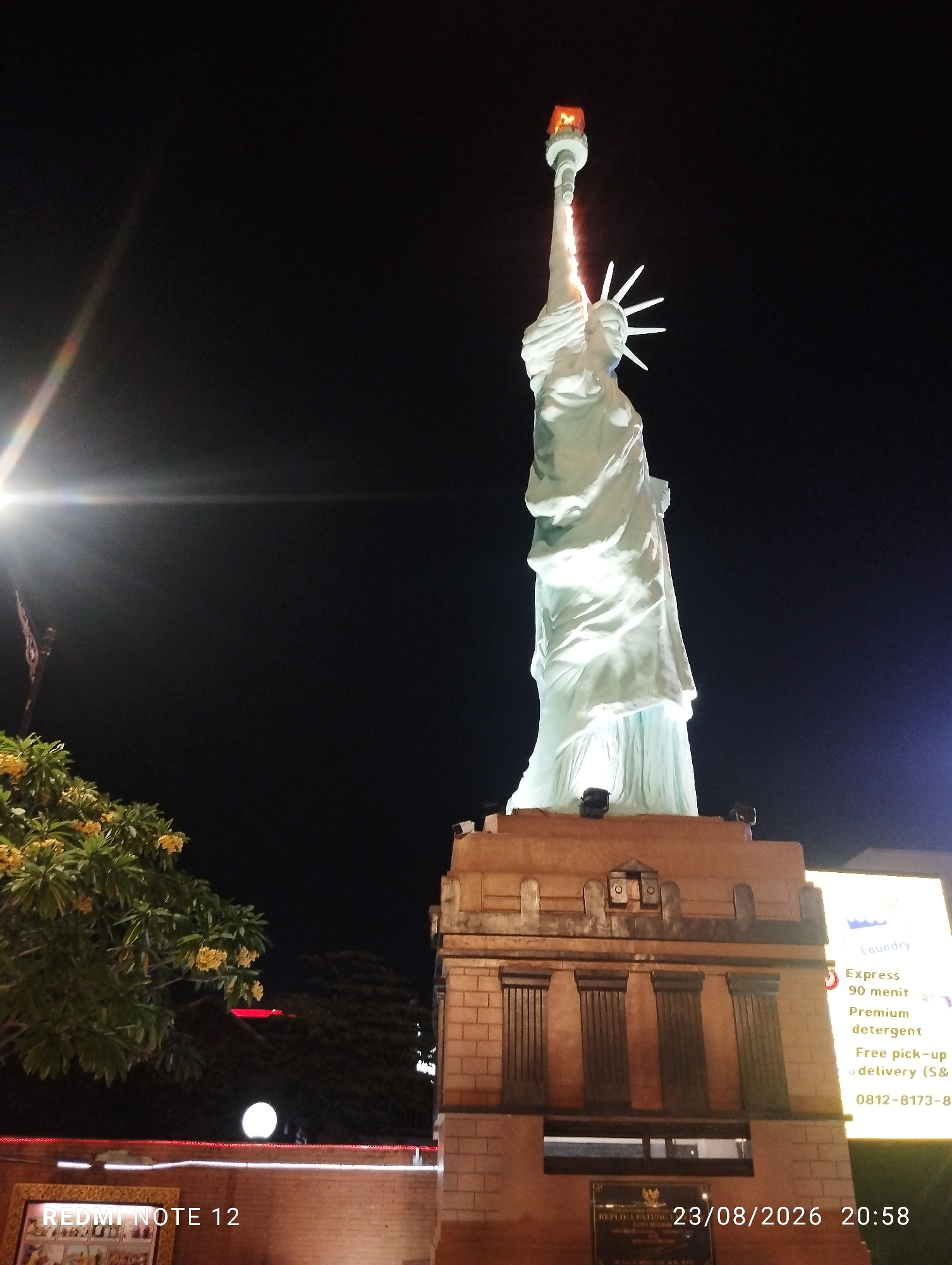 The image shows a nighttime exterior view of a replica of the Statue of Liberty, located in the city of Madiun, Indonesia. The statue is illuminated and stands on a large, stone pedestal. To the right of the statue, a brightly lit sign for a laundry service is visible. The sign advertises "Express 90 menit," "Premium detergent," and "Free pick-up & delivery (S&)." Below the laundry sign, a phone number "0812-8173-8..." is partially visible. On the base of the pedestal, a plaque with Indonesian text is present, along with the date and time "23/08/2026 20:58." In the lower left of the image, a framed artwork is partially visible, and the text "REDMI NOTE 12" is overlaid. A tree with yellow flowers is visible to the left of the statue. The sky is dark, and a bright light source is creating lens flare in the upper left quadrant of the image.