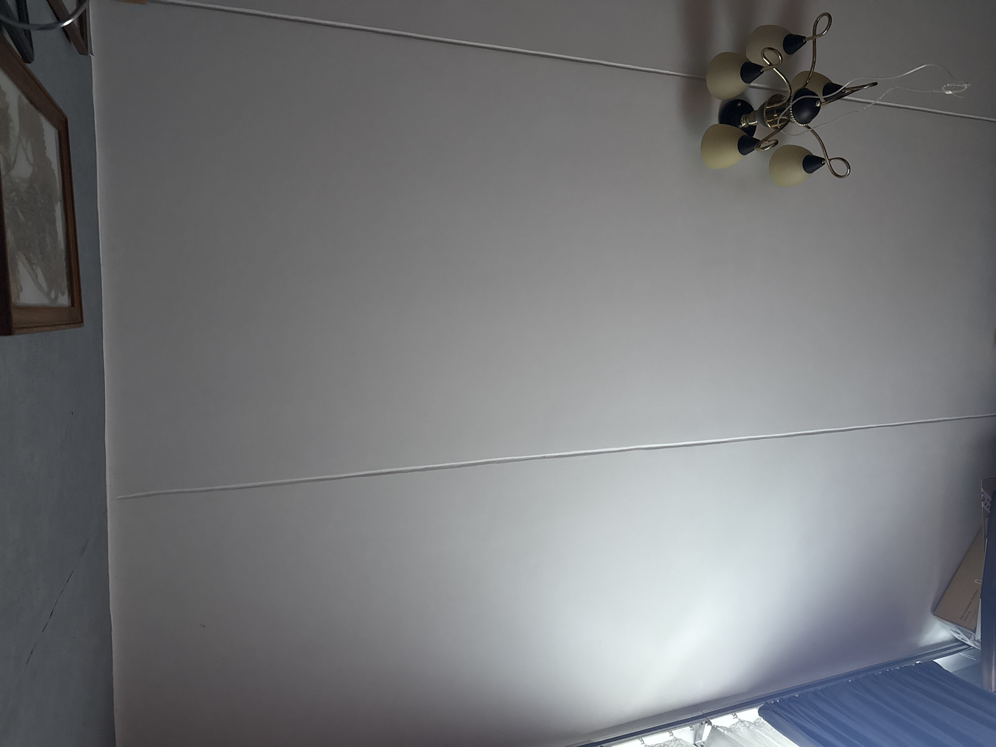 The image shows a ceiling with a light fixture. The ceiling appears to be paneled. The light fixture is a semi-flush mount with five frosted glass shades, each with a black base and brass accents. The fixture is suspended by a black stem. One of the glass shades is tilted. A white wire is hanging from the fixture. A framed picture with an image of what appears to be a map or a drawing is visible on the wall above the ceiling. There are also dark blue curtains visible on the left side of the image, and a cardboard box is partially visible on a surface below. The scene is indoors and appears to be a domestic setting. There is no indication of the location being Moscow, Russia.
