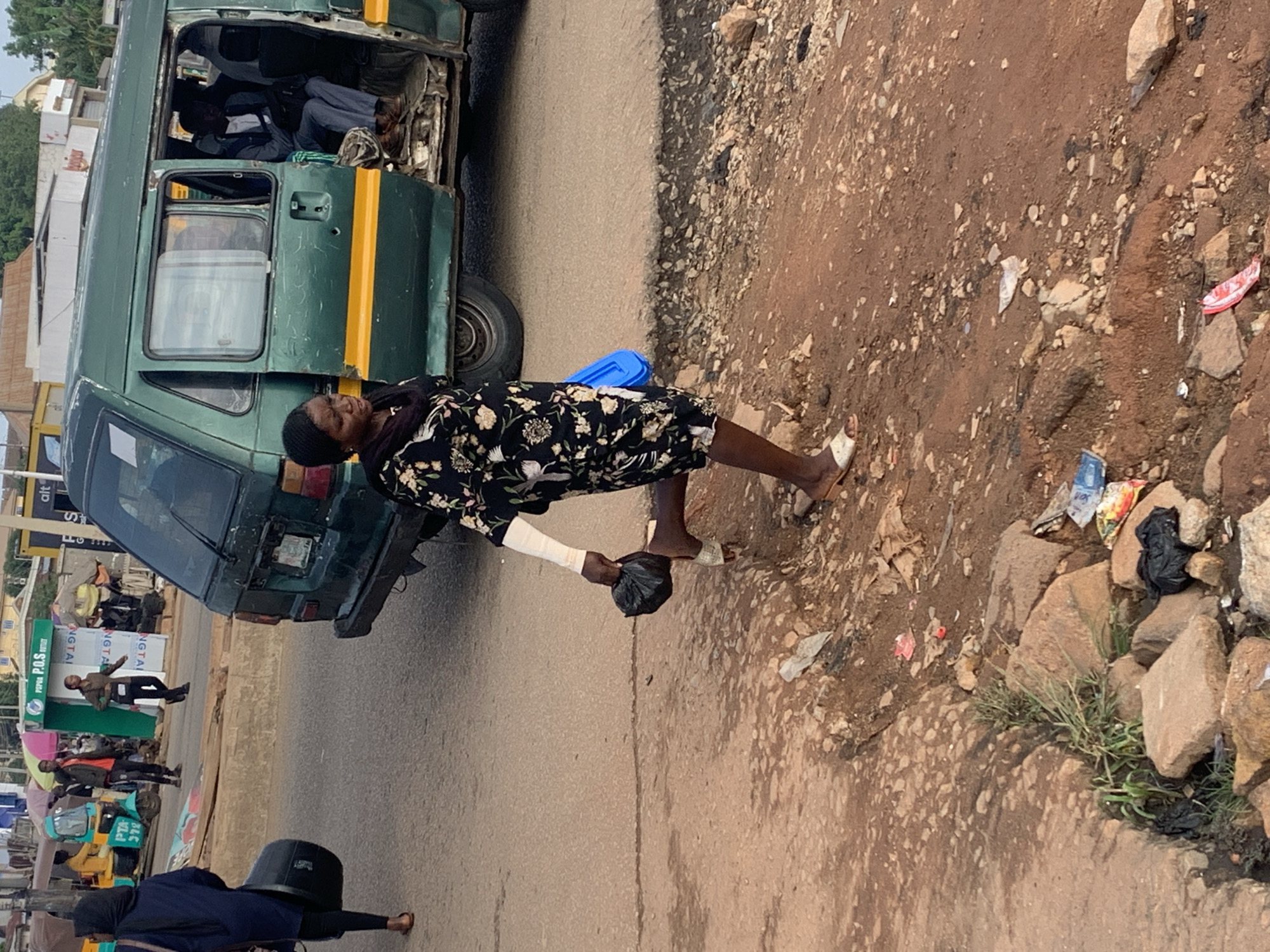 The image depicts a street scene in Gura Top, Nigeria, with a green minibús parked on the right side of the frame. Several people are visible, including a woman in the foreground walking away from the camera, carrying a black plastic bag in her right hand and a blue object in her left. She is wearing a dark floral-print dress and white sandals. Her left arm is bandaged. Behind her, a woman wearing a dark hijab is walking towards the camera, carrying a large black drum. Inside the minibús, several passengers are seated, one of whom is wearing a blue jacket and grey trousers. In the background, other individuals and a yellow and blue tricycle-like vehicle are present, suggesting a busy urban environment. The ground is a mix of asphalt and dirt, with visible debris, rocks, and patchy vegetation. The overall impression is that of a bustling, perhaps somewhat unkempt, public thoroughfare.