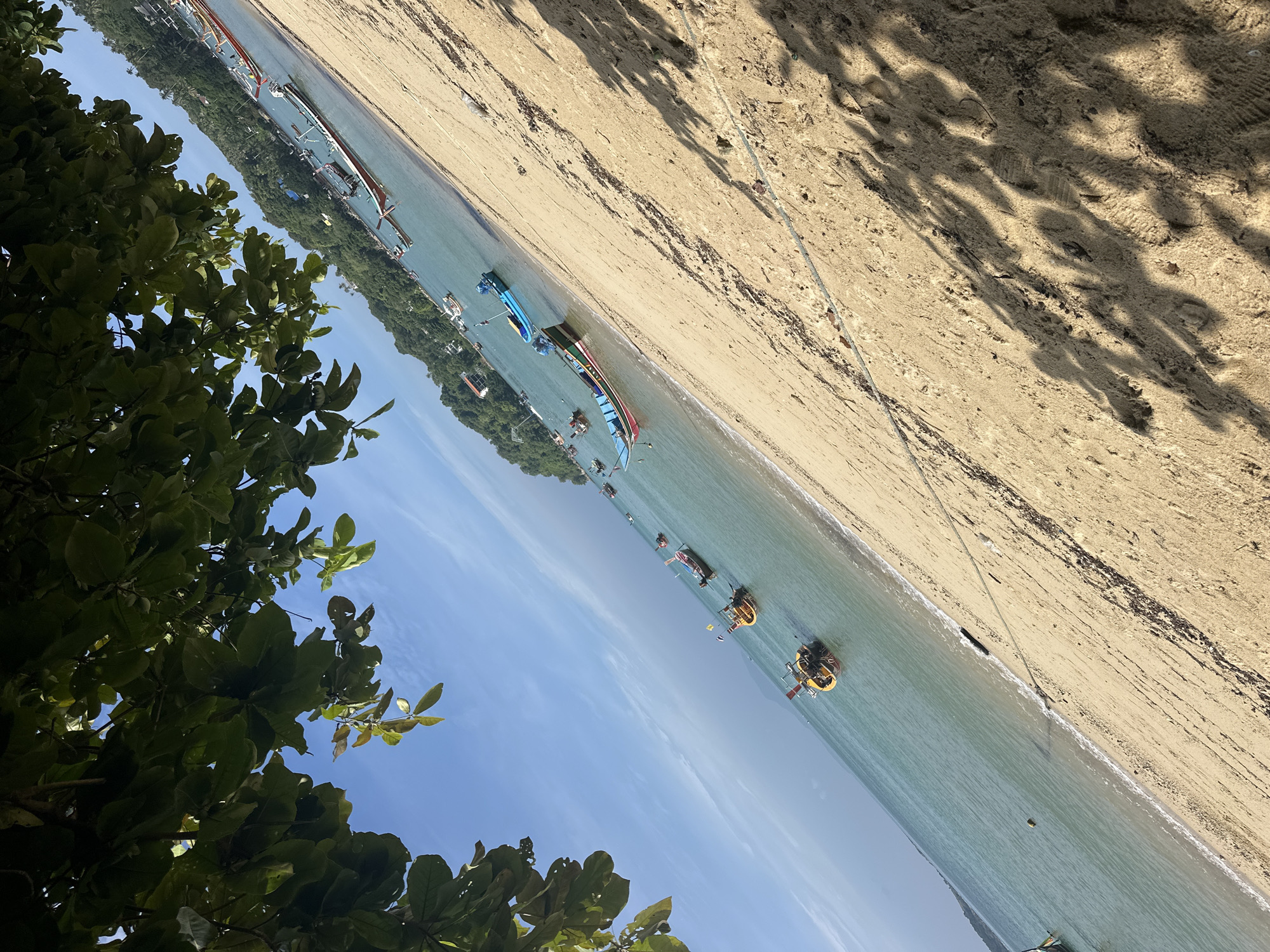The image captures a sunlit beach scene in Rawai, Thailand. The foreground is dominated by a sandy beach, with a thick rope stretched across it and shadows of foliage cast by unseen trees. The sea occupies the middle ground, appearing calm and clear with several traditional longtail boats moored in the turquoise water. These boats, varying in color and size, some with yellow and red accents, are characteristic of Thai fishing vessels. In the background, a verdant, tree-covered hillside rises, dotted with a few buildings, indicating a coastal settlement. The sky is a bright blue with wispy white clouds. The composition is angled, giving a sense of depth and a diagonal perspective. There are no people visible in the frame.
