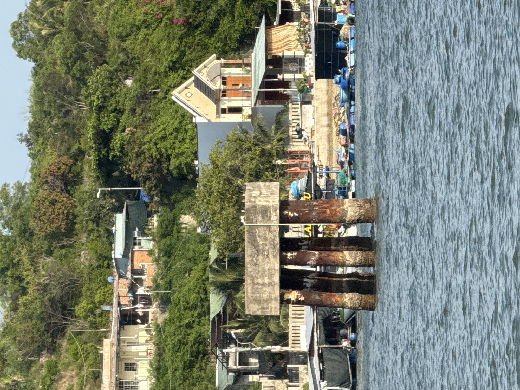 The image depicts a coastal scene in Nha Trang Ward, Vietnam, characterized by a lush green hillside densely covered with trees and vegetation. In the foreground, the calm, blue water of the sea gently ripples. Prominently in the middle ground is a weathered concrete structure supported by four thick, rusty metal pilings that rise from the water.
Along the shoreline, a row of buildings is visible, nestled against the hillside. These structures vary in architectural style and condition, with some appearing to be houses and others more utilitarian buildings, possibly associated with the waterfront activities. Several boats are moored or floating near the shore, some of which are small fishing vessels or rafts, indicated by nets and blue barrels.
Further back on the hillside, more buildings are scattered among the trees, suggesting a residential area. The overall impression is one of a developing coastal community, with a mix of natural landscape and human habitation. There are no visible people in the image, and no specific actions or interactions are depicted. The environment is sunny, with clear skies.