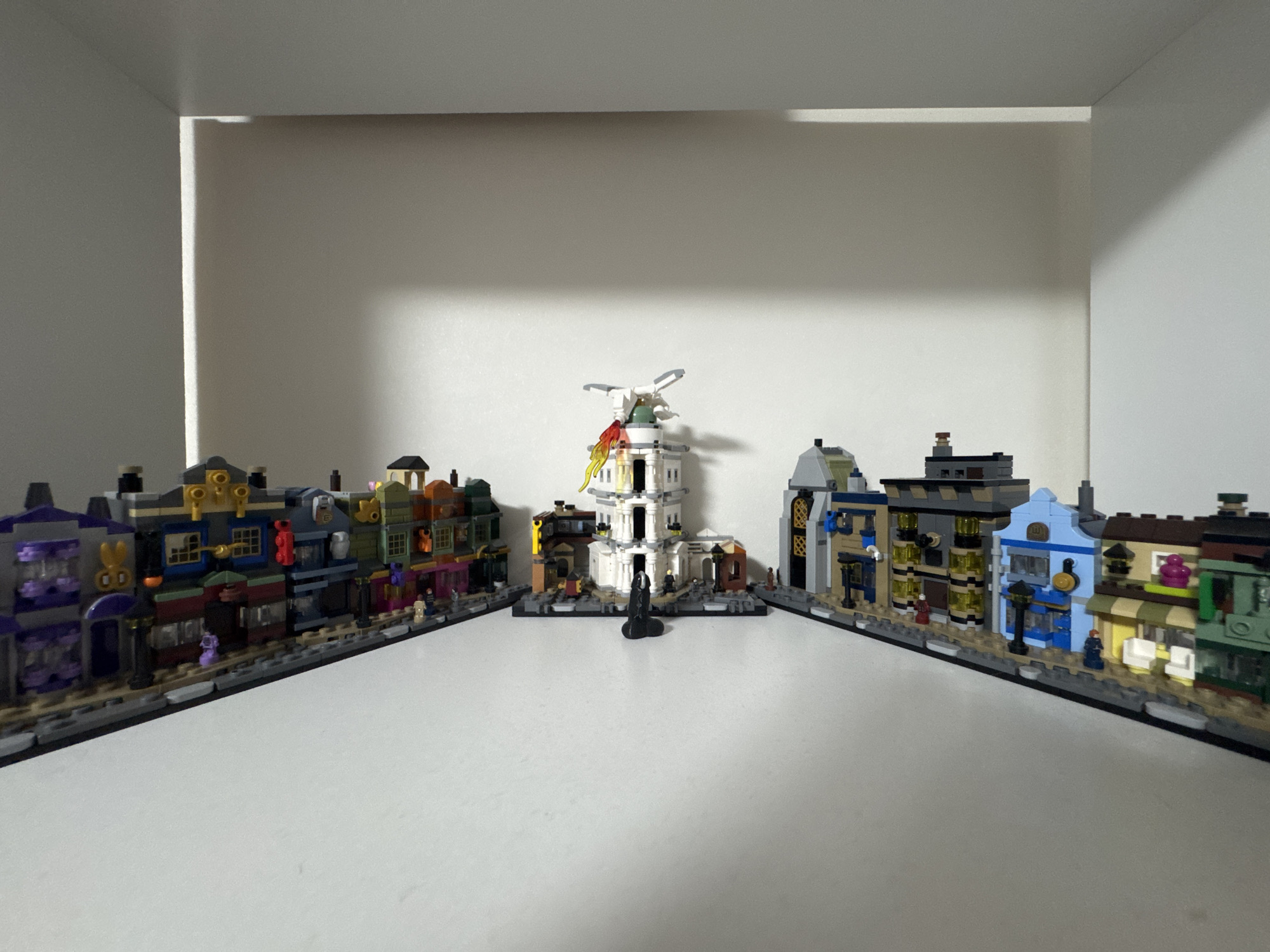 The image shows a collection of LEGO buildings arranged on a white shelf. In the center is a white tower with a windmill on top, emitting yellow and red flame-like pieces. Flanking the tower are two rows of LEGO shops, one on the left and one on the right. The shops are colorful and detailed, featuring various architectural styles and small LEGO figures. The environment appears to be indoors, likely a room with a white shelving unit. There is no discernible connection to the city of Kyiv, Ukraine, or any specific real-world location.