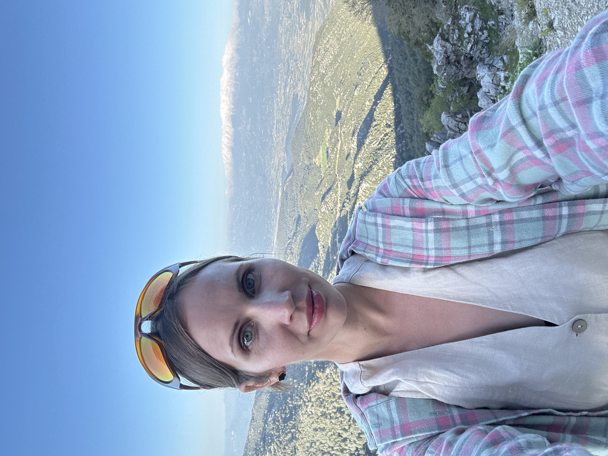 A woman is taking a selfie. She is wearing a plaid shirt and sunglasses on her head. The background shows a landscape including mountains, trees, and city. The location is likely Fethiye, Turkey.