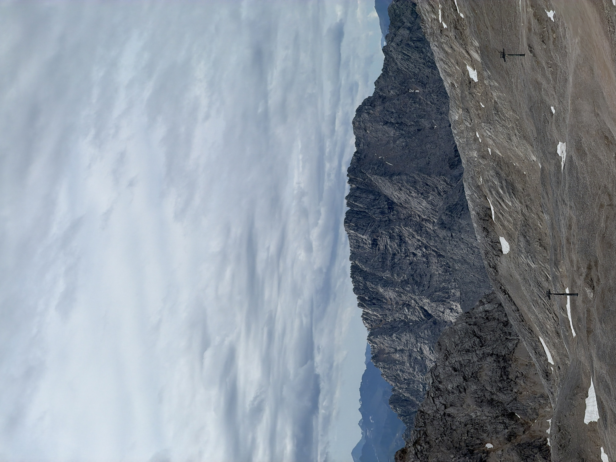The image depicts a vast, rugged mountain landscape under a cloudy sky. The foreground is characterized by a barren, rocky terrain with patches of snow. Two dark, vertical poles, likely ski lift towers, are visible in the mid-ground. In the background, a massive, jagged mountain range dominates the horizon, its rocky slopes conveying a sense of immense scale and geological age. The overall atmosphere is one of wilderness and solitude. This scene is consistent with the summit area of the Zugspitze, Germany's highest peak, located near Garmisch-Partenkirchen.